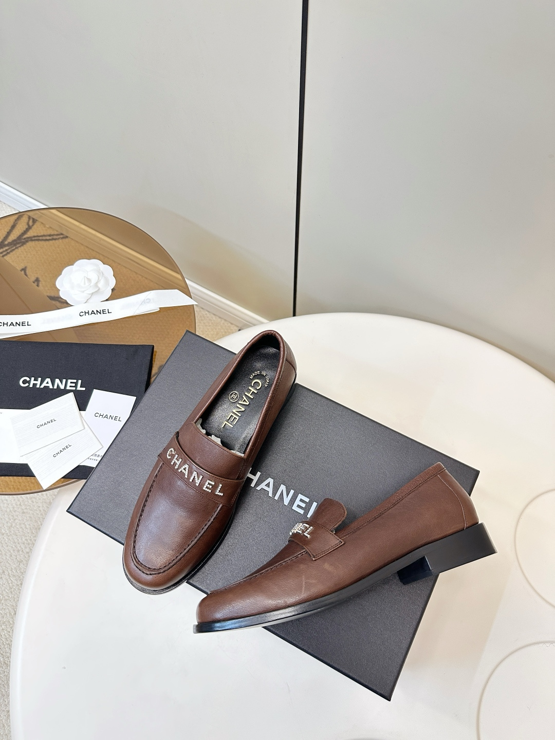 A pair of brown Chanel leather loafers with crystal logo lettering on the strap, presented on a black Chanel box with accessories. - Hình 9