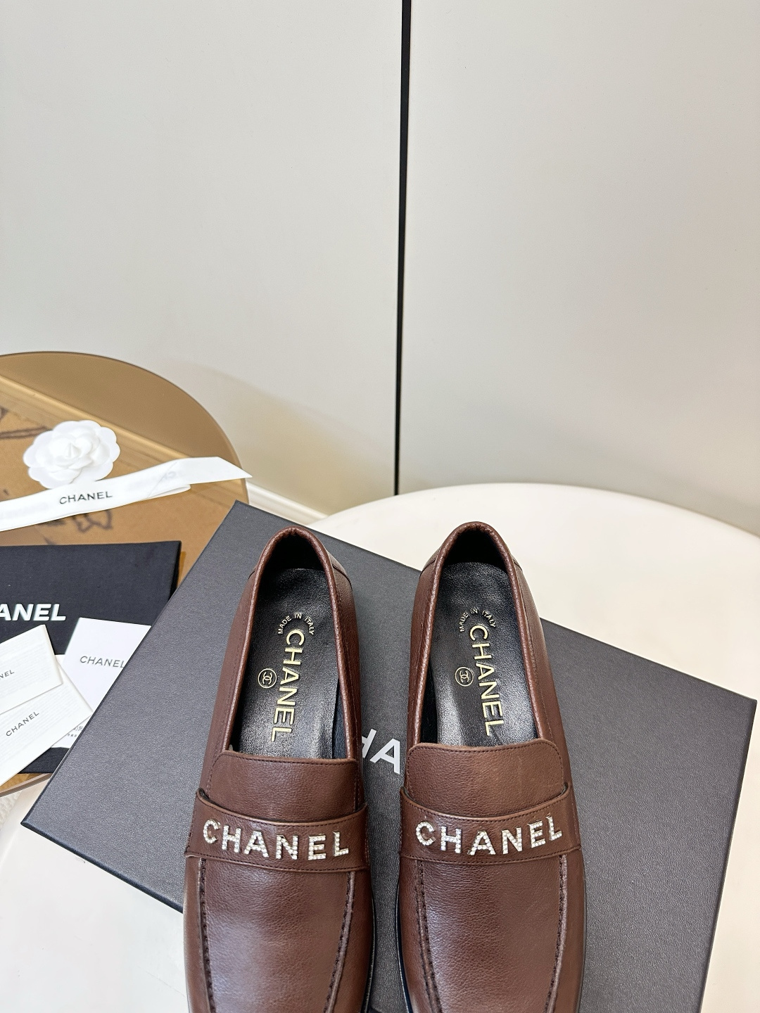 A pair of brown Chanel leather loafers with crystal logo lettering on the strap, presented on a black Chanel box with accessories. - Hình 8