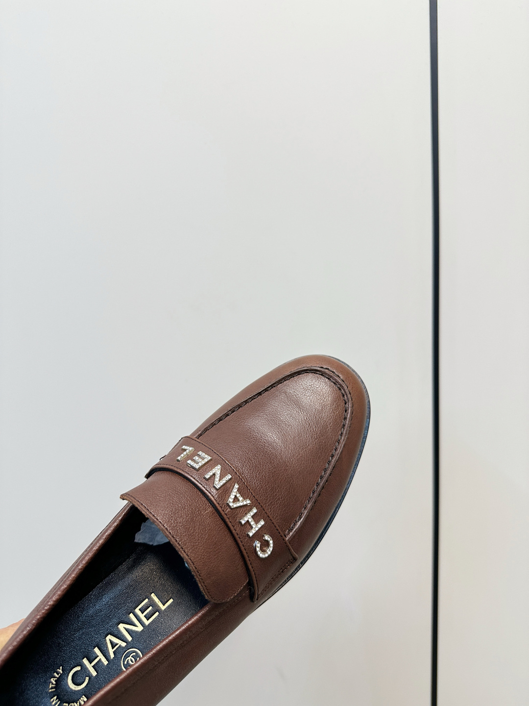 A pair of brown Chanel leather loafers with crystal logo lettering on the strap, presented on a black Chanel box with accessories. - Hình 7