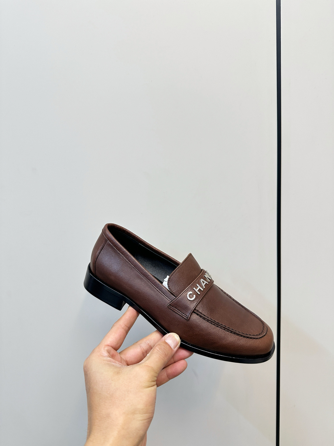 A pair of brown Chanel leather loafers with crystal logo lettering on the strap, presented on a black Chanel box with accessories. - Hình 6