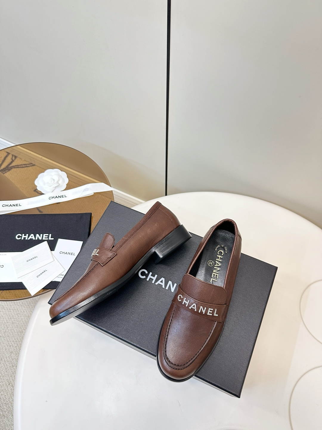 A pair of brown Chanel leather loafers with crystal logo lettering on the strap, presented on a black Chanel box with accessories. - Hình 5