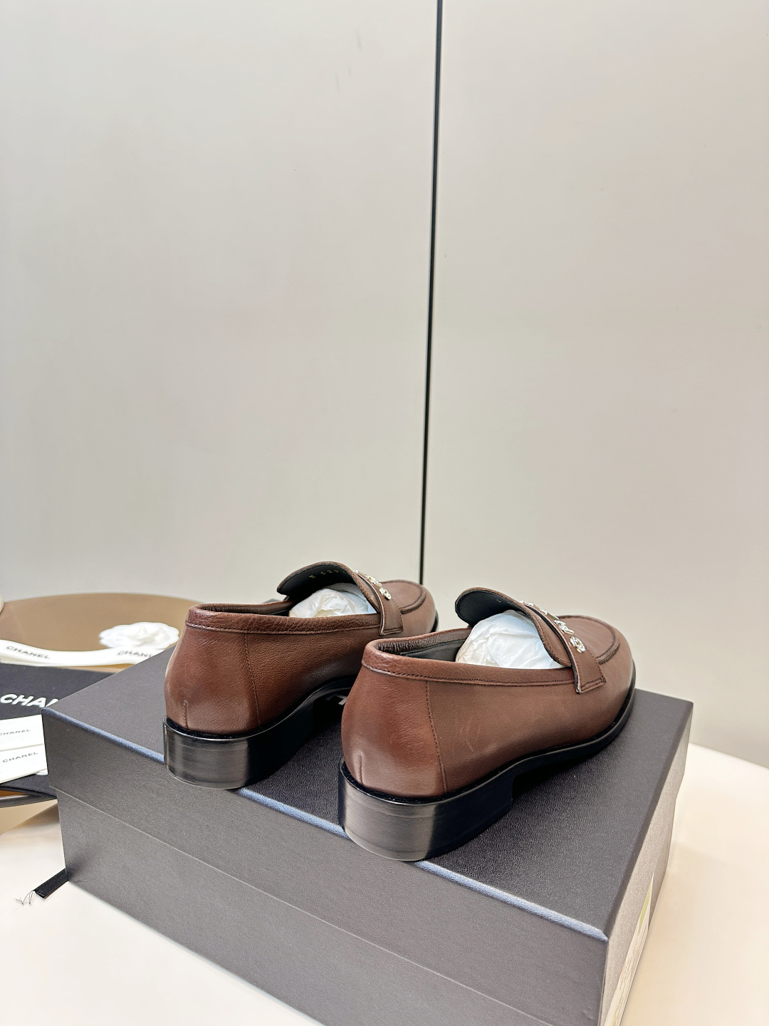 A pair of brown Chanel leather loafers with crystal logo lettering on the strap, presented on a black Chanel box with accessories. - Hình 4