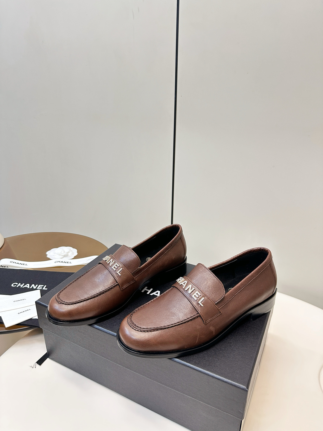 A pair of brown Chanel leather loafers with crystal logo lettering on the strap, presented on a black Chanel box with accessories. - Hình 3