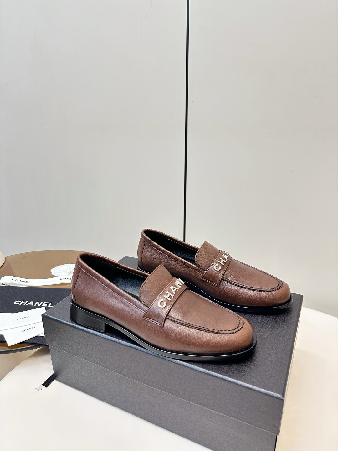 A pair of brown Chanel leather loafers with crystal logo lettering on the strap, presented on a black Chanel box with accessories. - Hình 2