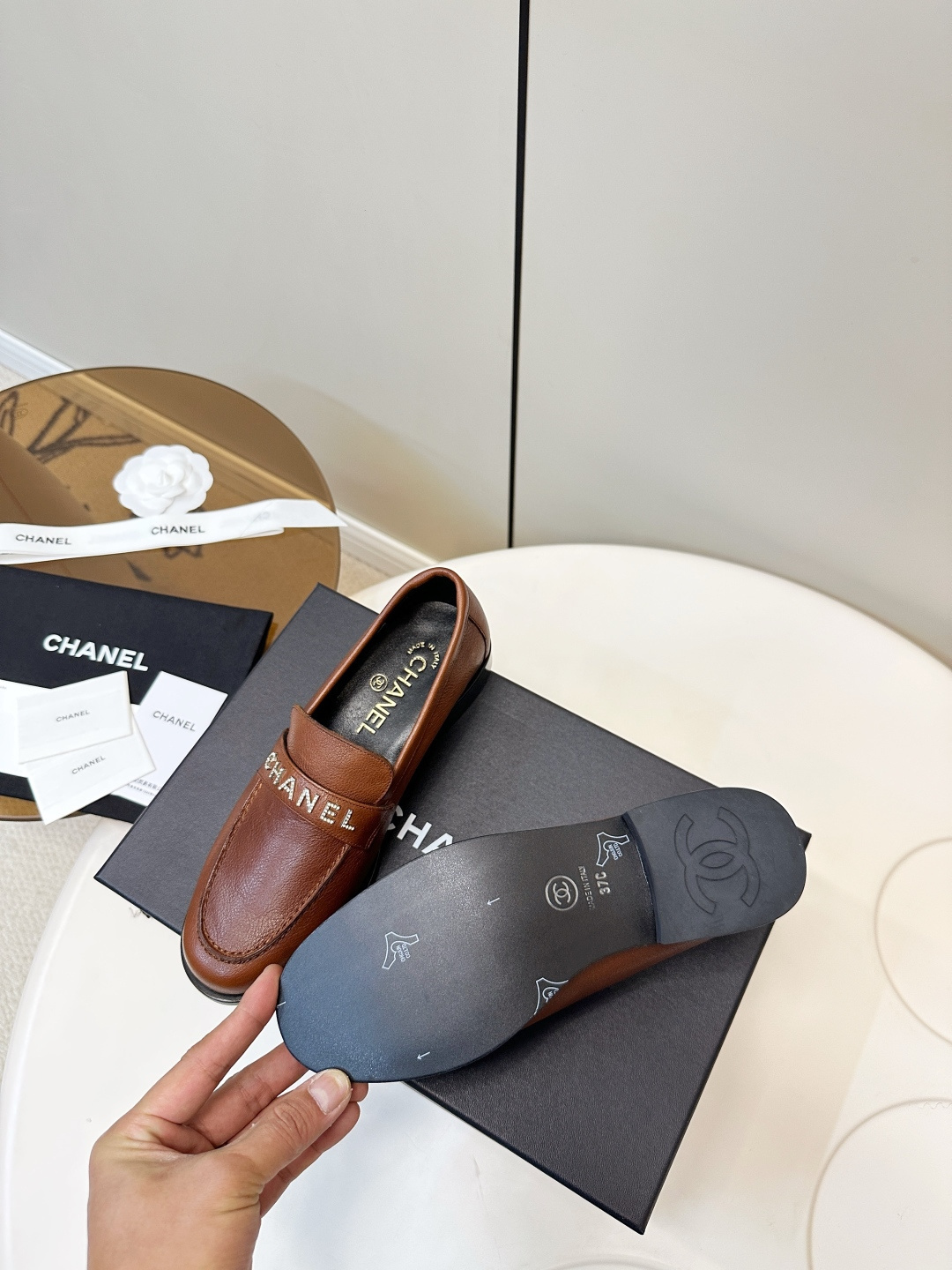 Elegant Chanel brown leather loafers with crystal-embellished logo displayed on a black designer box. - Hình 10