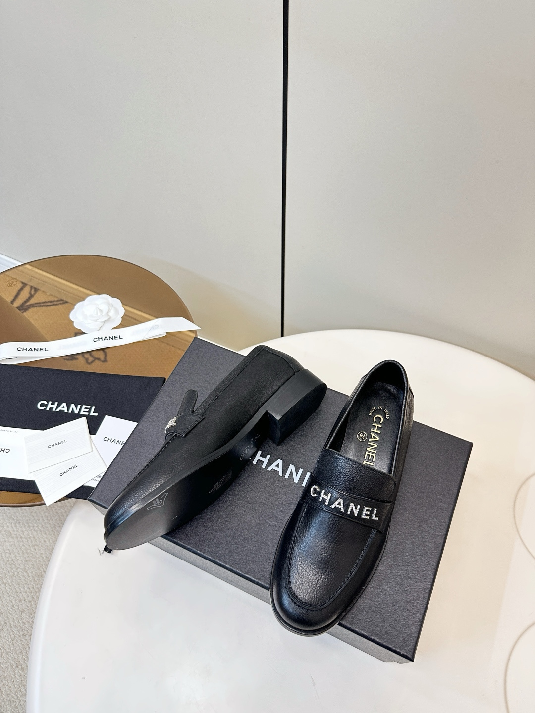 A pair of black Chanel leather loafers with silver crystal logo embellishment displayed on a black box. - Hình 5