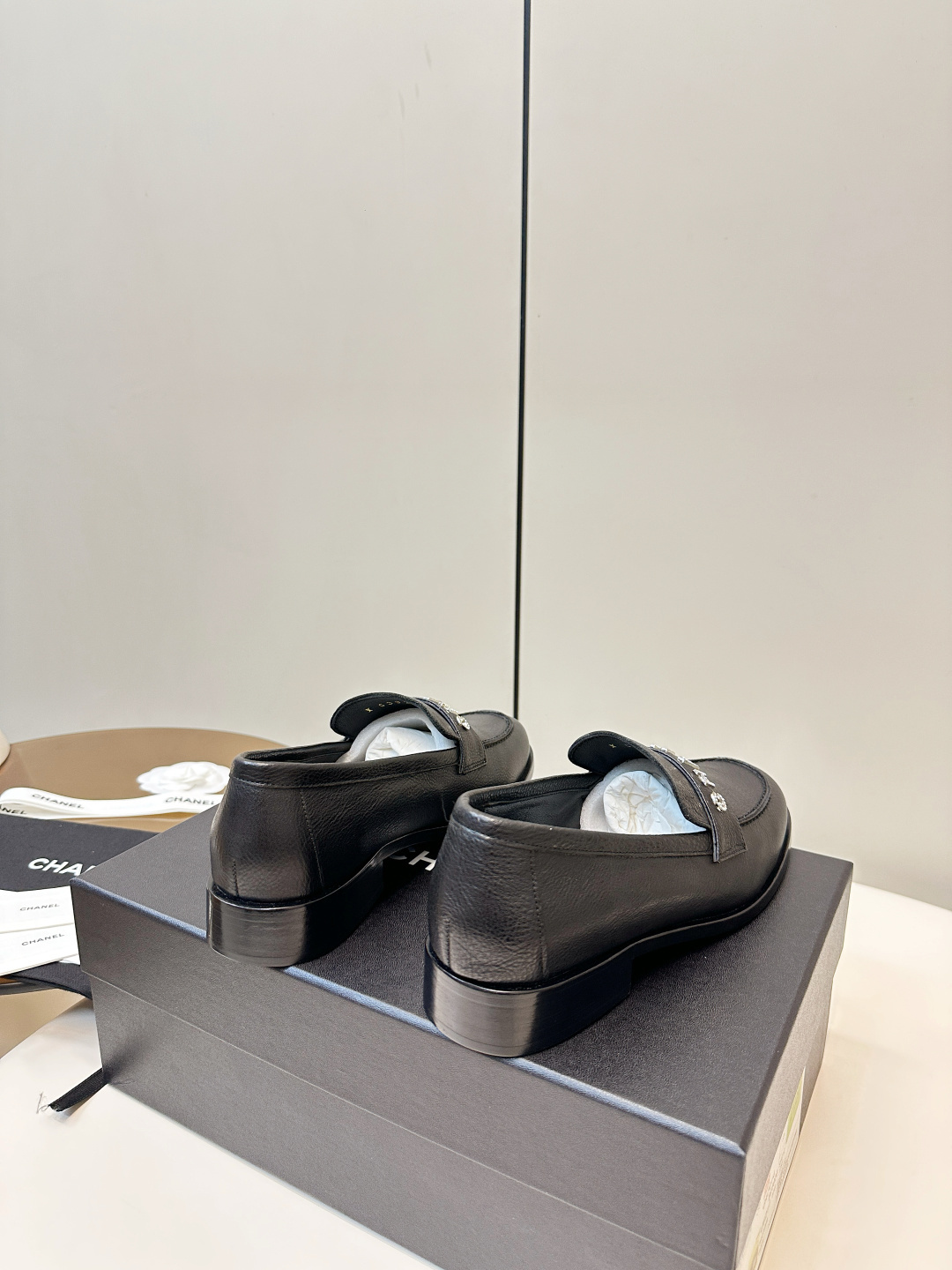 A pair of black Chanel leather loafers with silver crystal logo embellishment displayed on a black box. - Hình 4