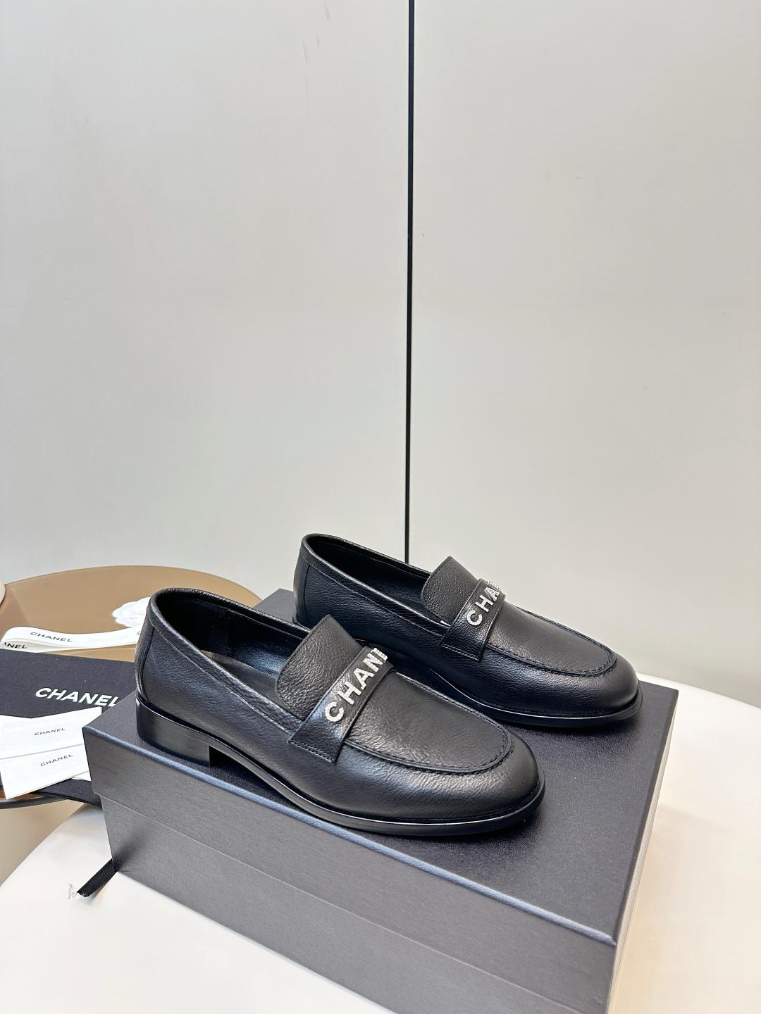 A pair of black Chanel leather loafers with silver crystal logo embellishment displayed on a black box. - Hình 2
