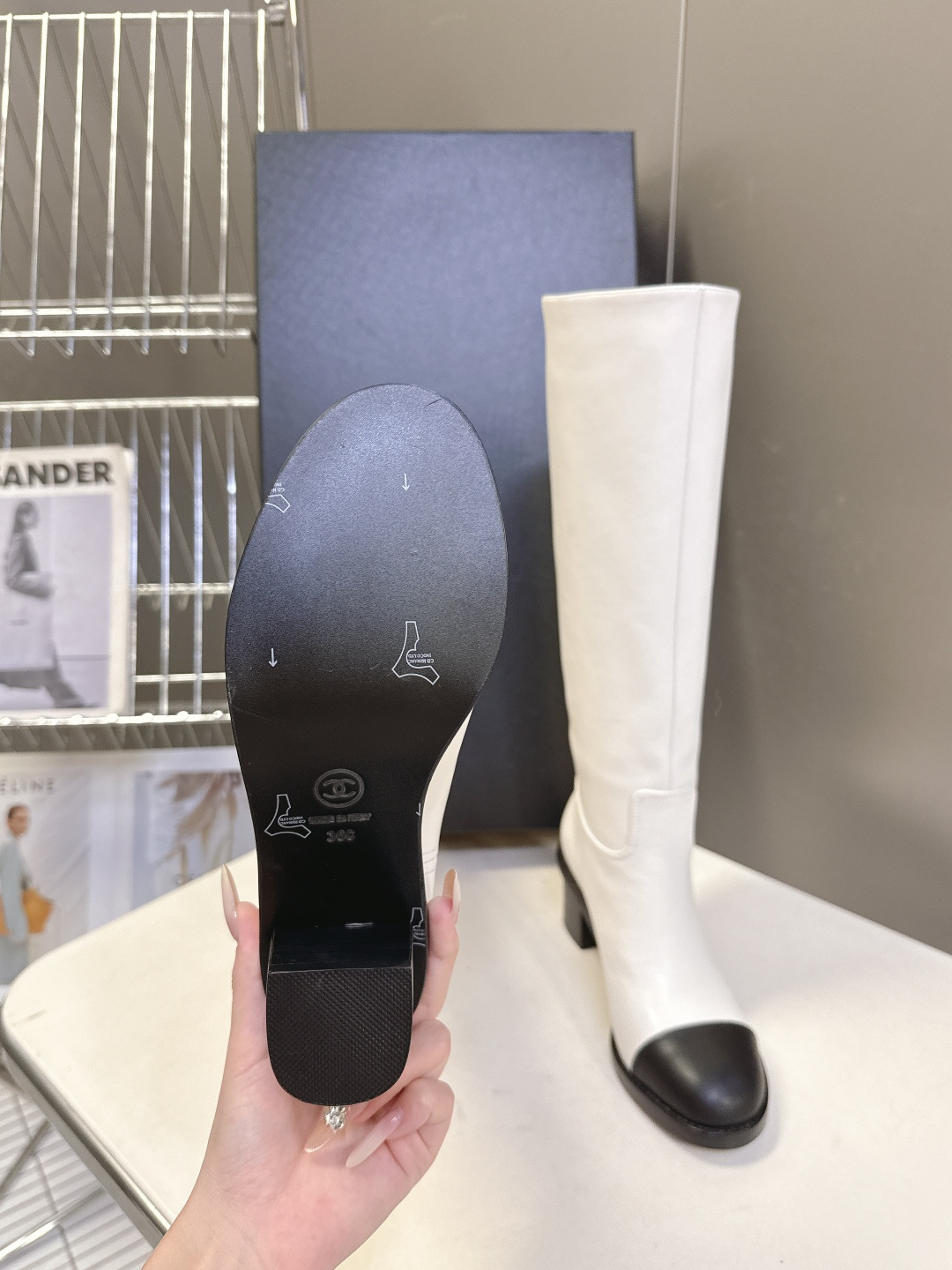 Elegant white leather knee-high boots with black cap toe and embroidered logo displayed with a designer box. - Hình 9