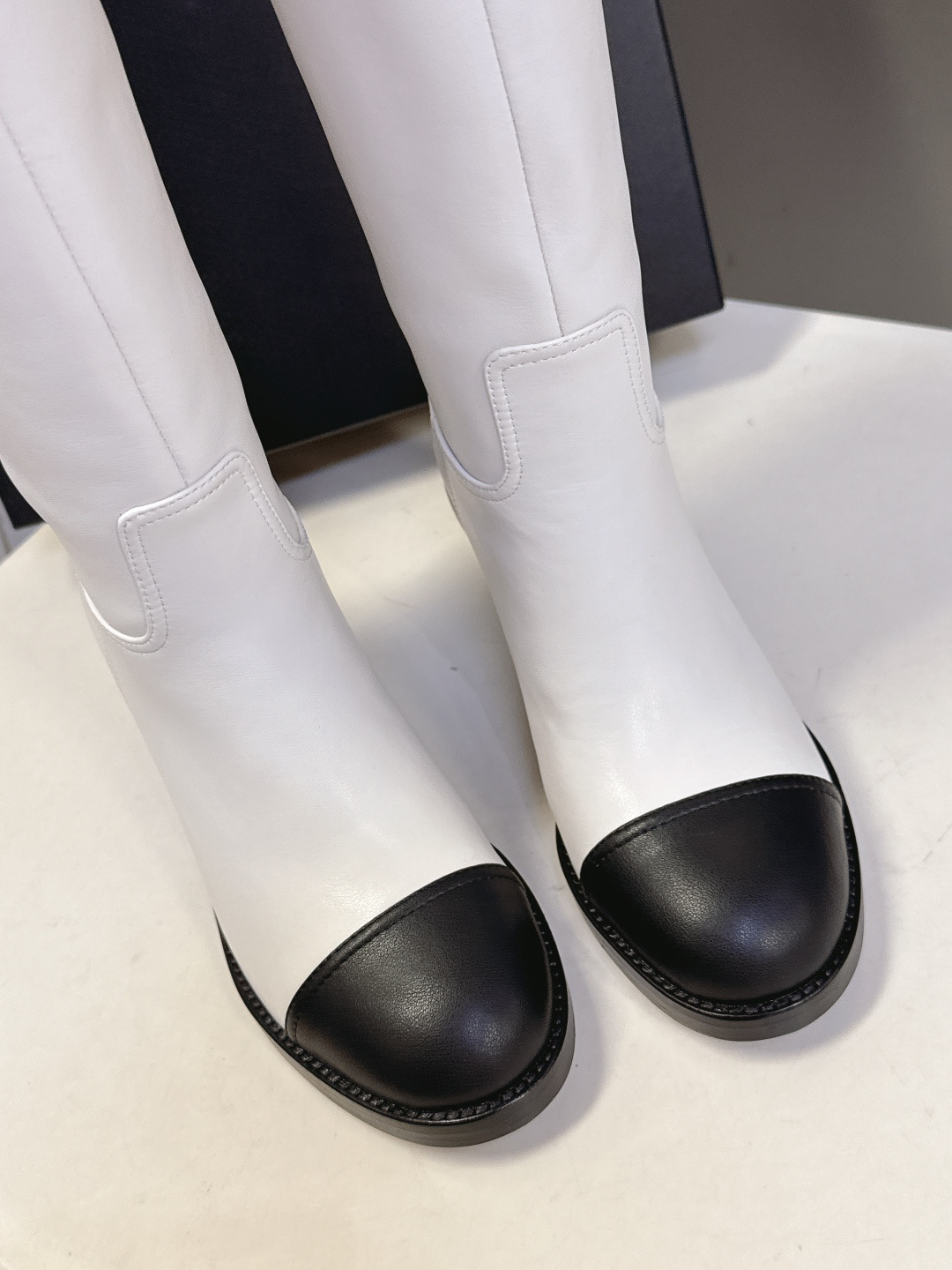 Elegant white leather knee-high boots with black cap toe and embroidered logo displayed with a designer box. - Hình 7