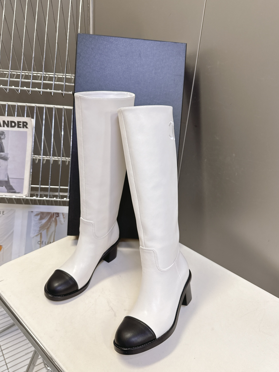 Elegant white leather knee-high boots with black cap toe and embroidered logo displayed with a designer box. - Hình 4