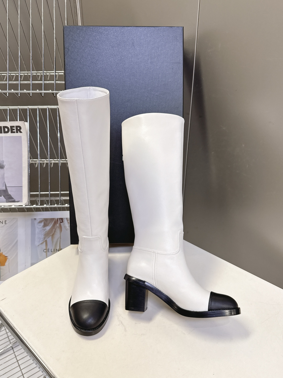 Elegant white leather knee-high boots with black cap toe and embroidered logo displayed with a designer box. - Hình 3