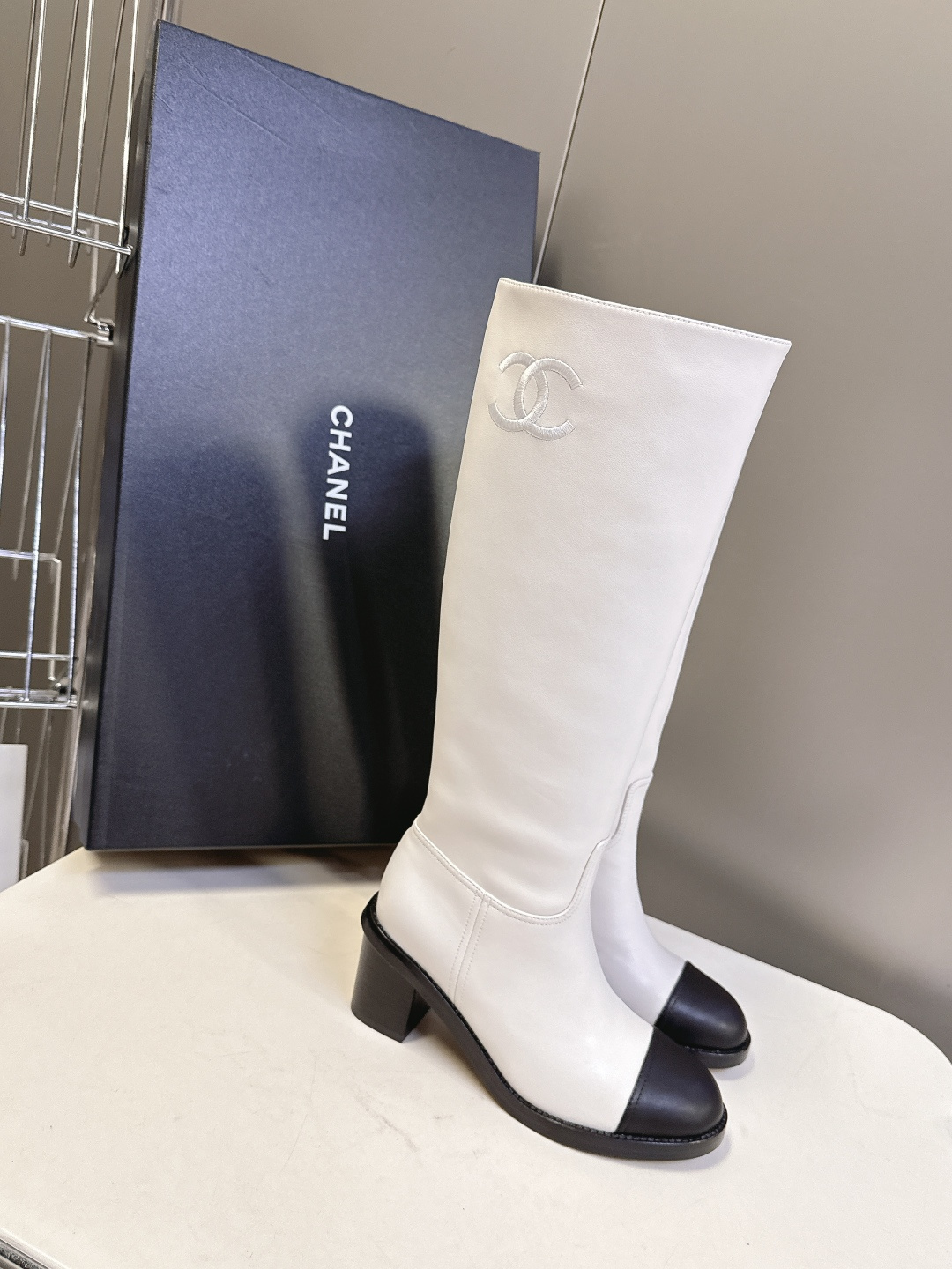Elegant white leather knee-high boots with black cap toe and embroidered logo displayed with a designer box. - Hình 2