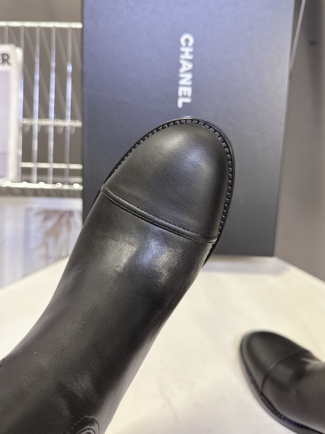 Pair of Chanel black leather knee-high boots with CC logo and block heel next to a Chanel shoe box - Hình 7