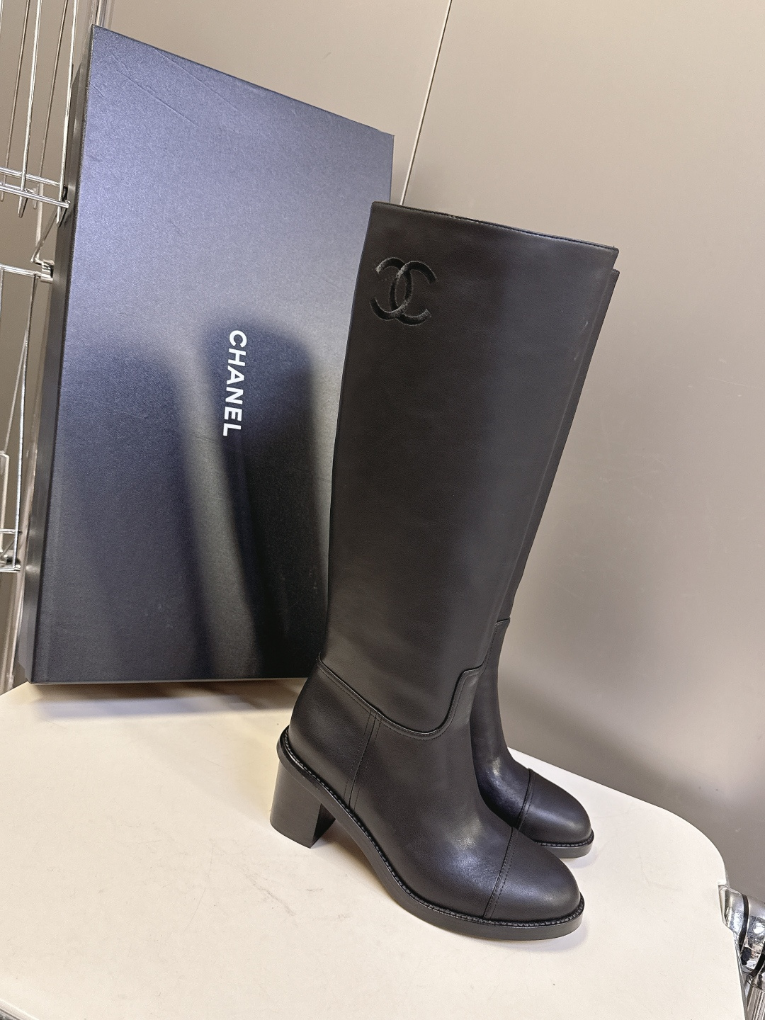 Pair of Chanel black leather knee-high boots with CC logo and block heel next to a Chanel shoe box - Hình 2