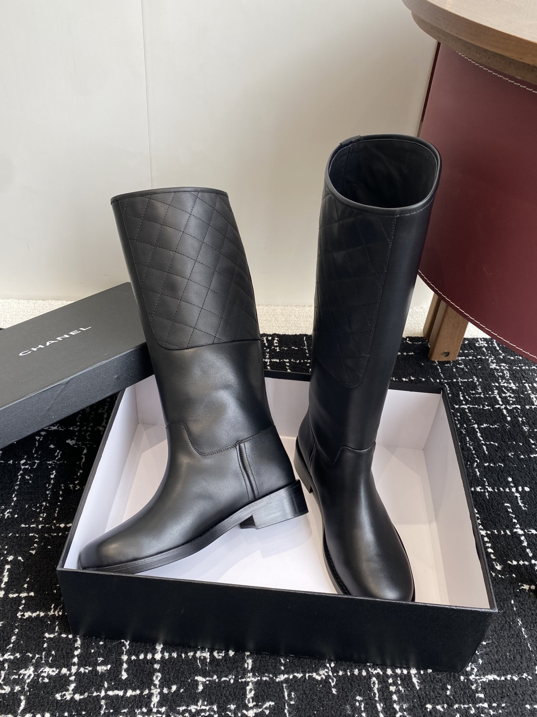 A pair of luxury Chanel black quilted leather knee-high riding boots placed on a black designer box. - Hình 5