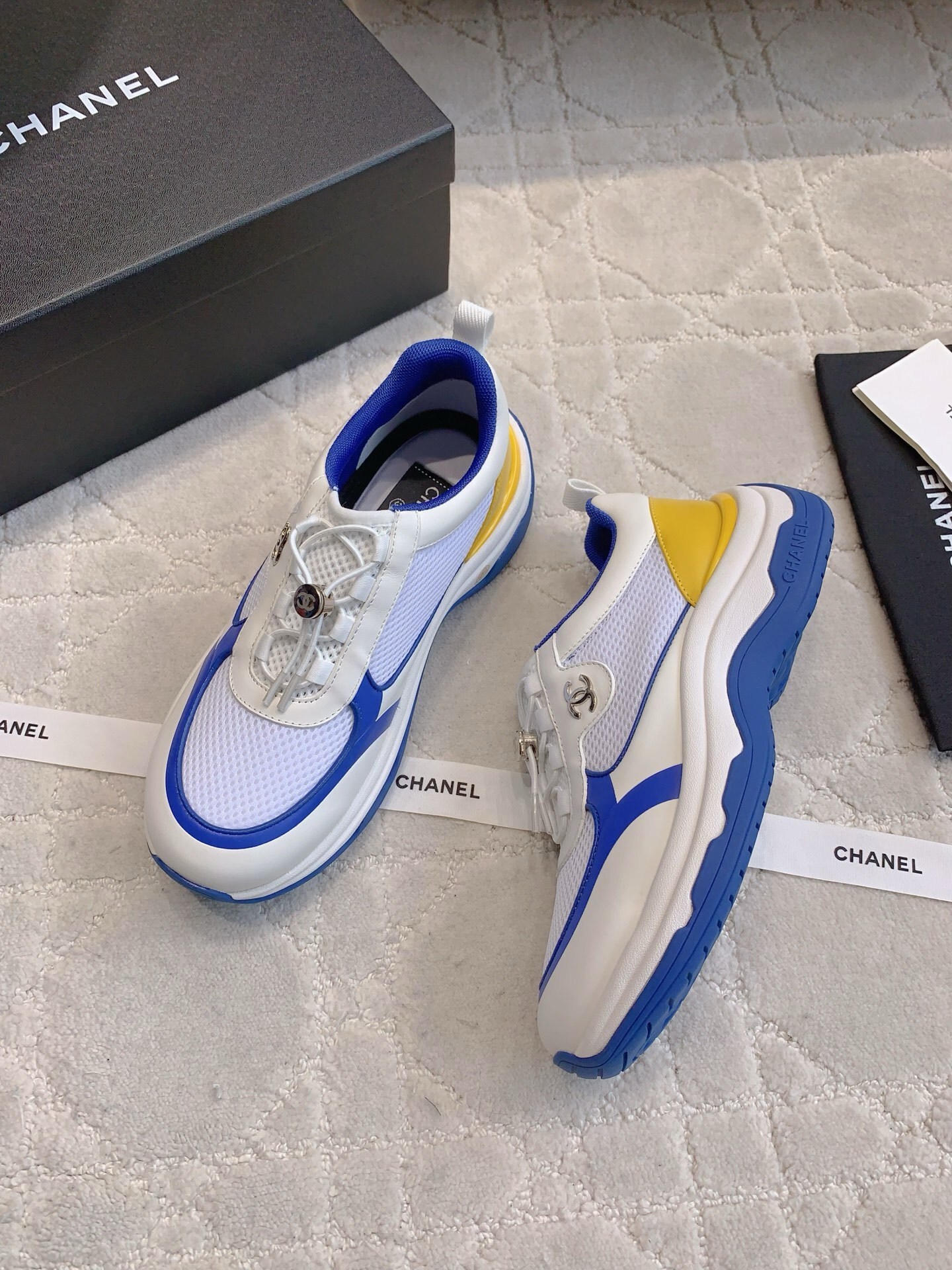 Chanel blue, white, and yellow mesh sneakers with CC logo toggle laces displayed with original packaging box and ribbons. - Hình 3