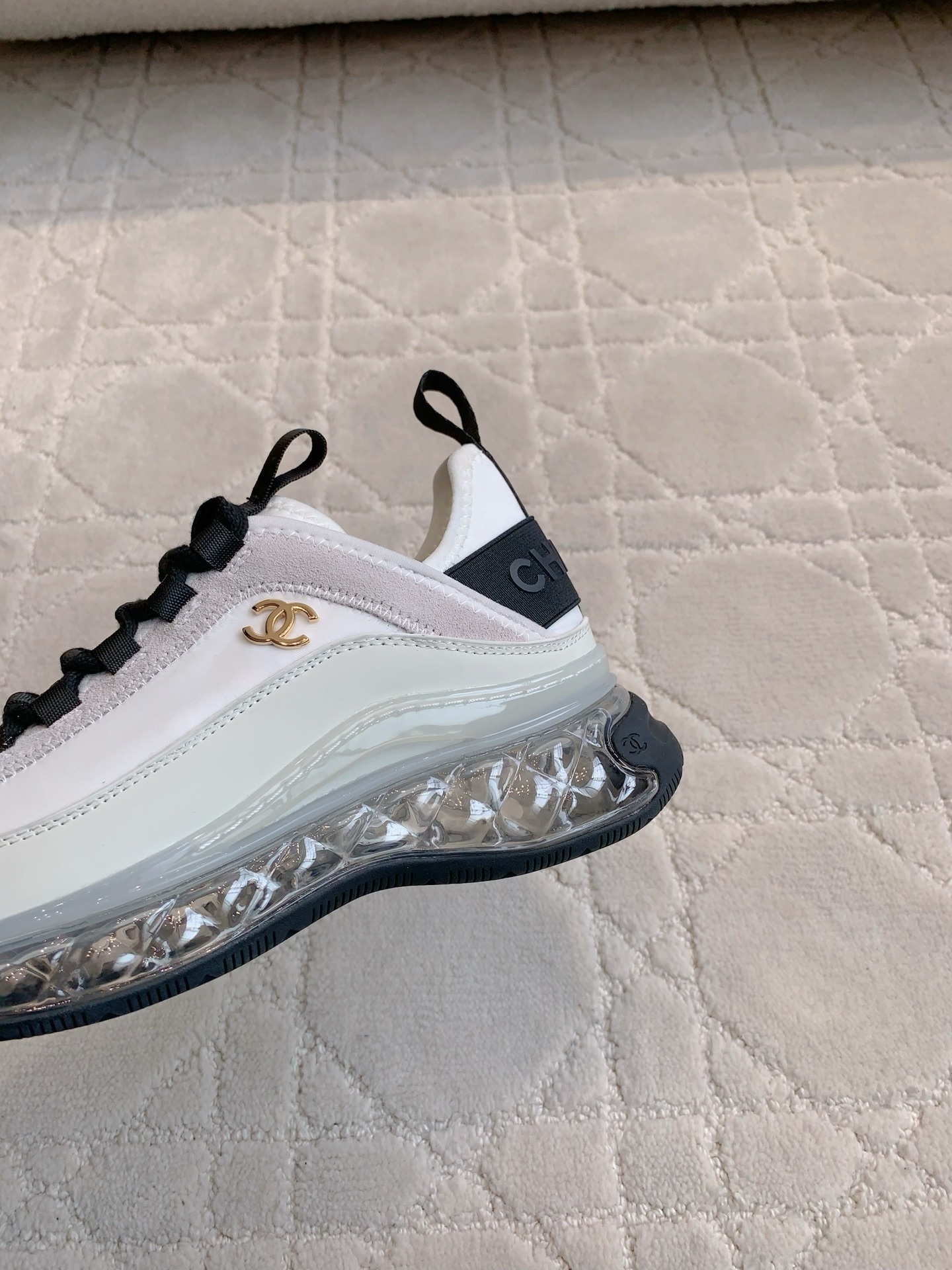 A pair of luxury Chanel white and black sneakers with transparent bubble soles and gold CC logo on a patterned carpet. - Hình 7