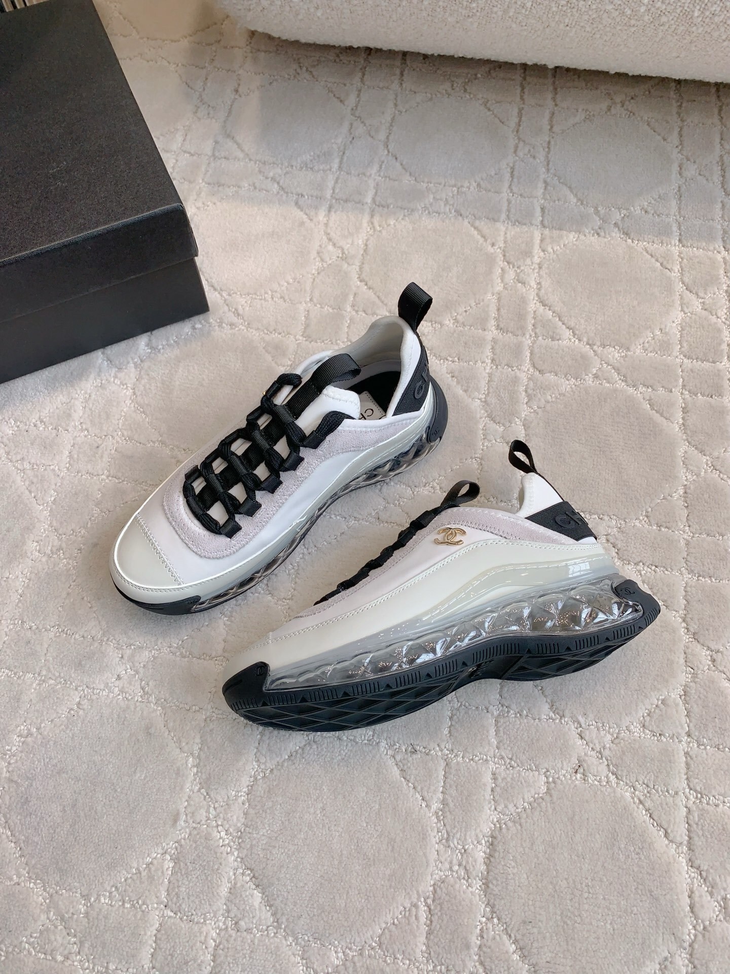 A pair of luxury Chanel white and black sneakers with transparent bubble soles and gold CC logo on a patterned carpet. - Hình 4