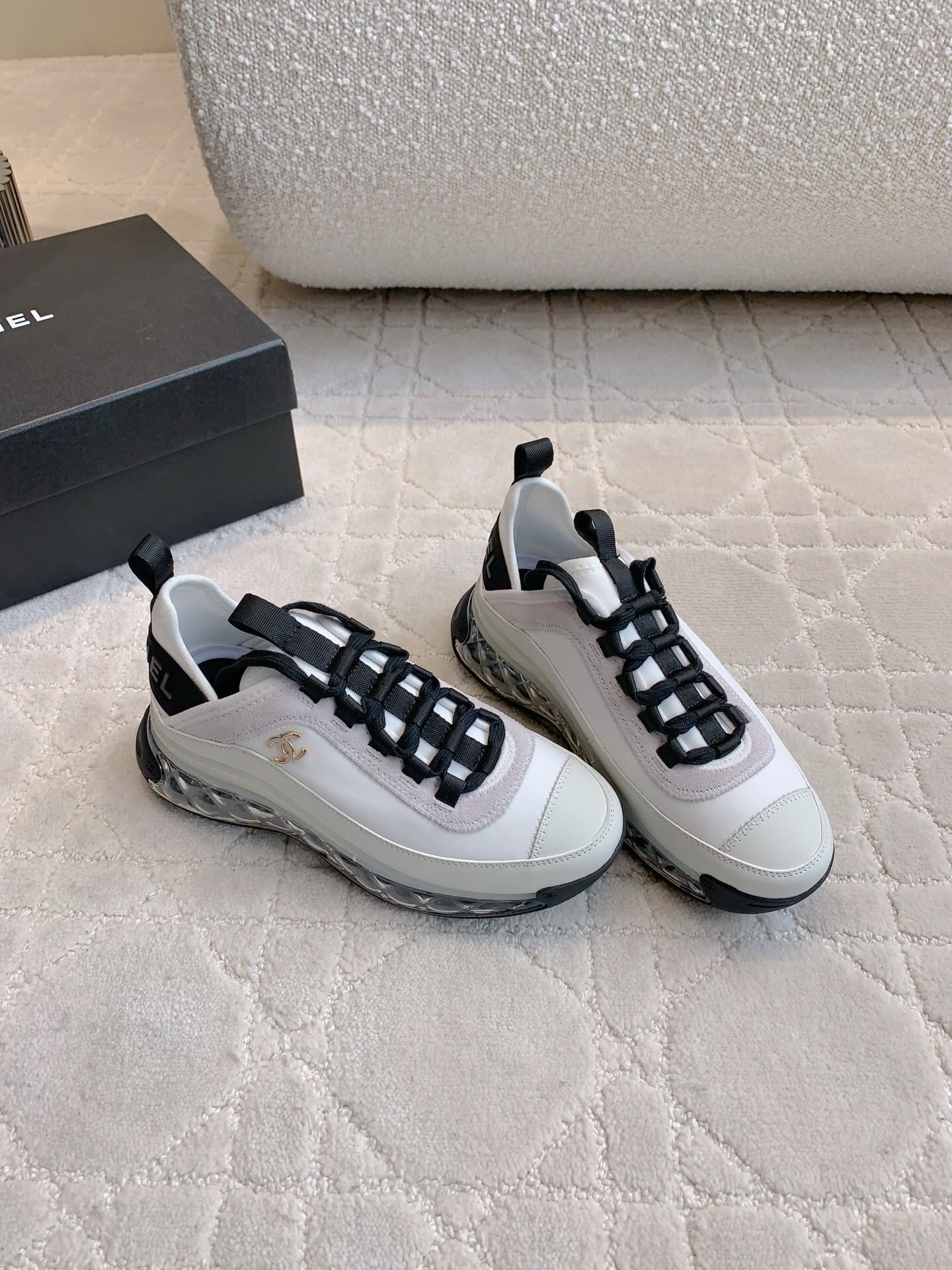 A pair of luxury Chanel white and black sneakers with transparent bubble soles and gold CC logo on a patterned carpet. - Hình 3