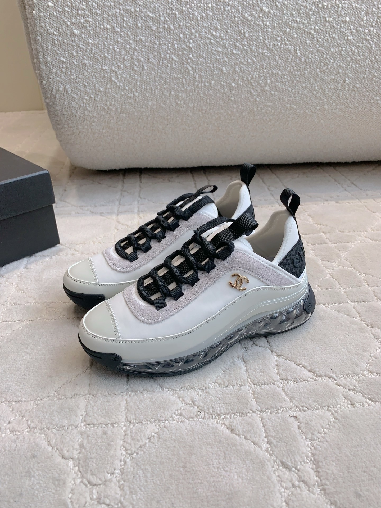 A pair of luxury Chanel white and black sneakers with transparent bubble soles and gold CC logo on a patterned carpet. - Hình 2