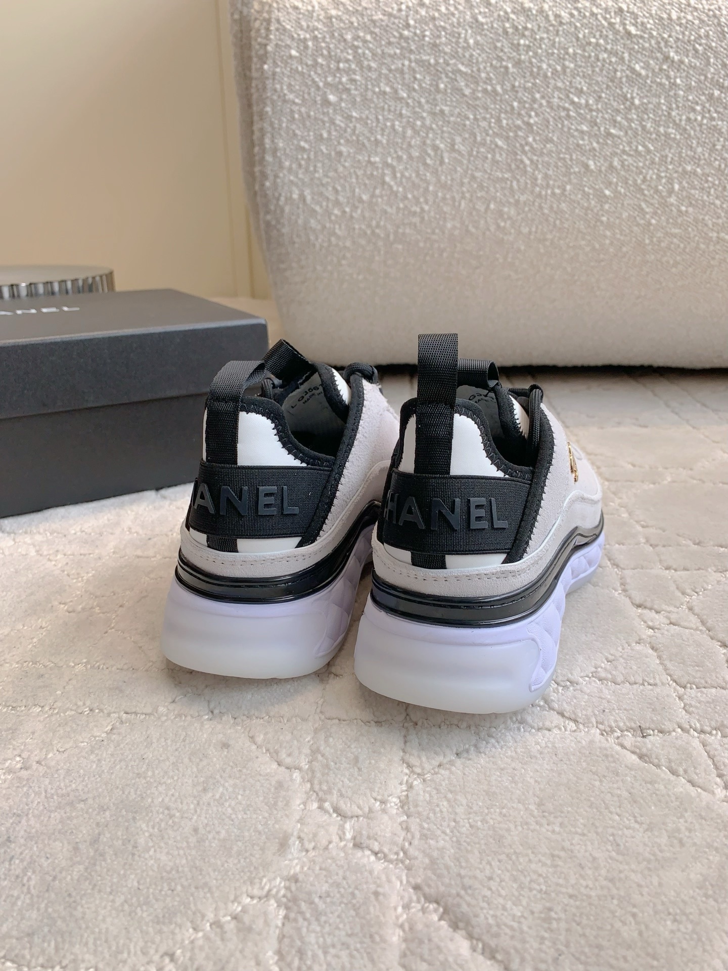 A pair of luxury Chanel white suede sneakers with black accents and gold CC logo placed on a cream quilted carpet. - Hình 8