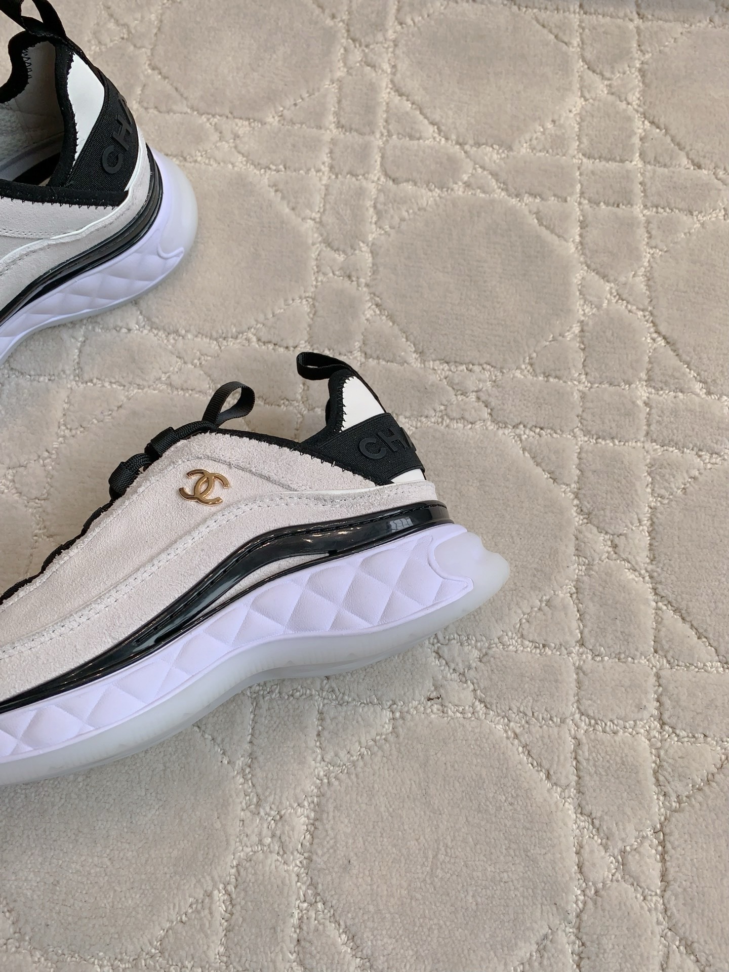 A pair of luxury Chanel white suede sneakers with black accents and gold CC logo placed on a cream quilted carpet. - Hình 7