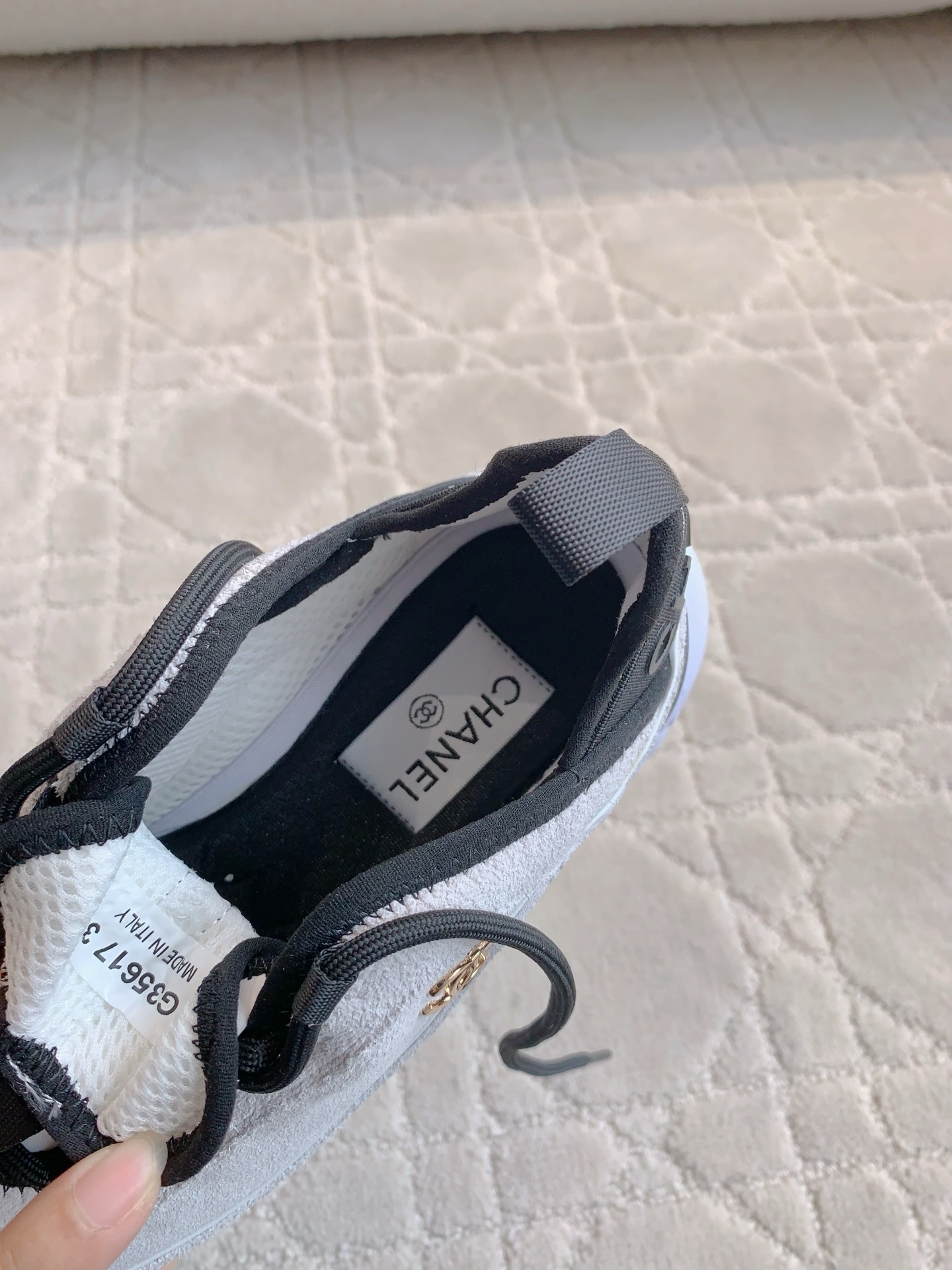 A pair of luxury Chanel white suede sneakers with black accents and gold CC logo placed on a cream quilted carpet. - Hình 6