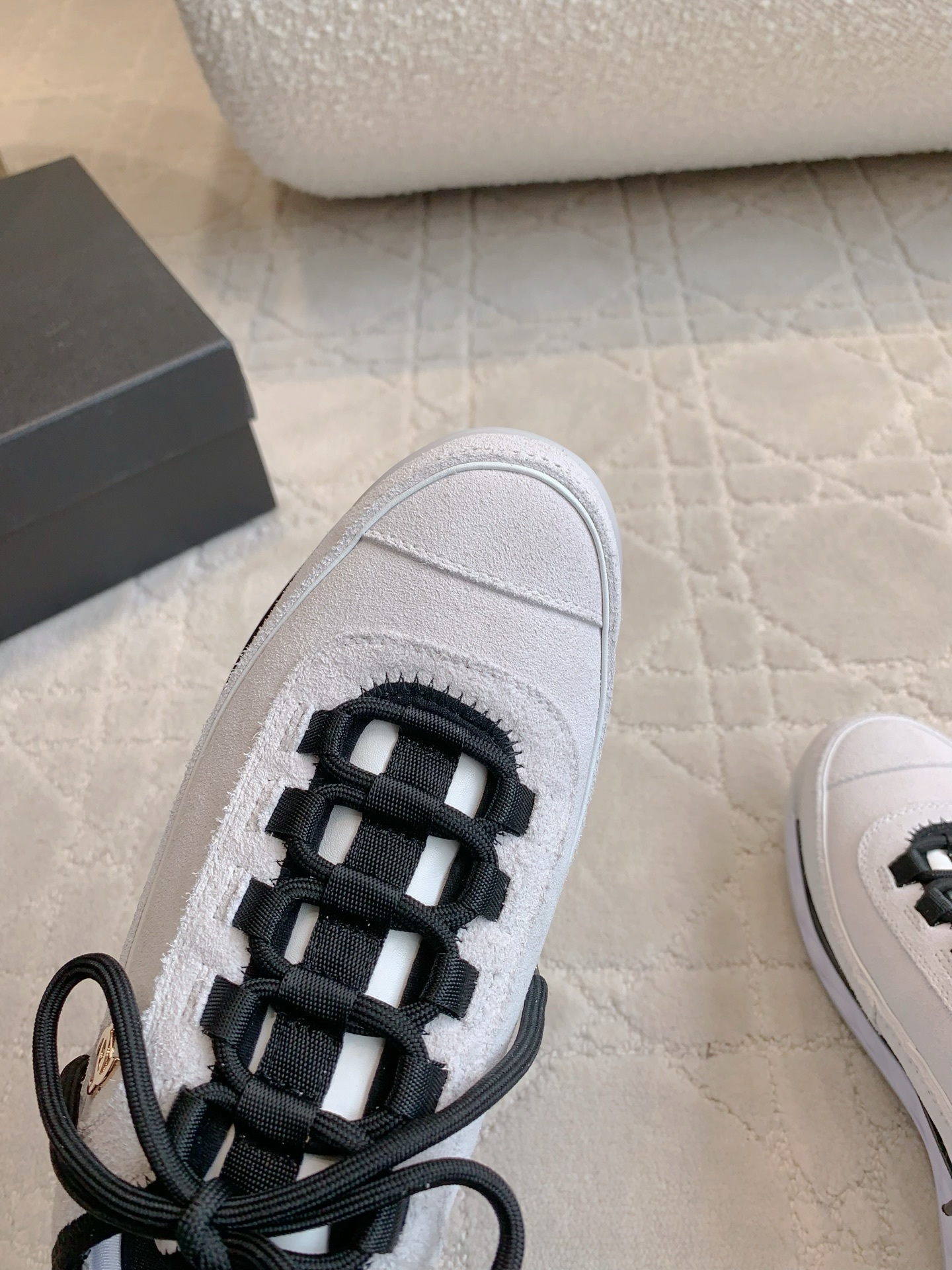 A pair of luxury Chanel white suede sneakers with black accents and gold CC logo placed on a cream quilted carpet. - Hình 5
