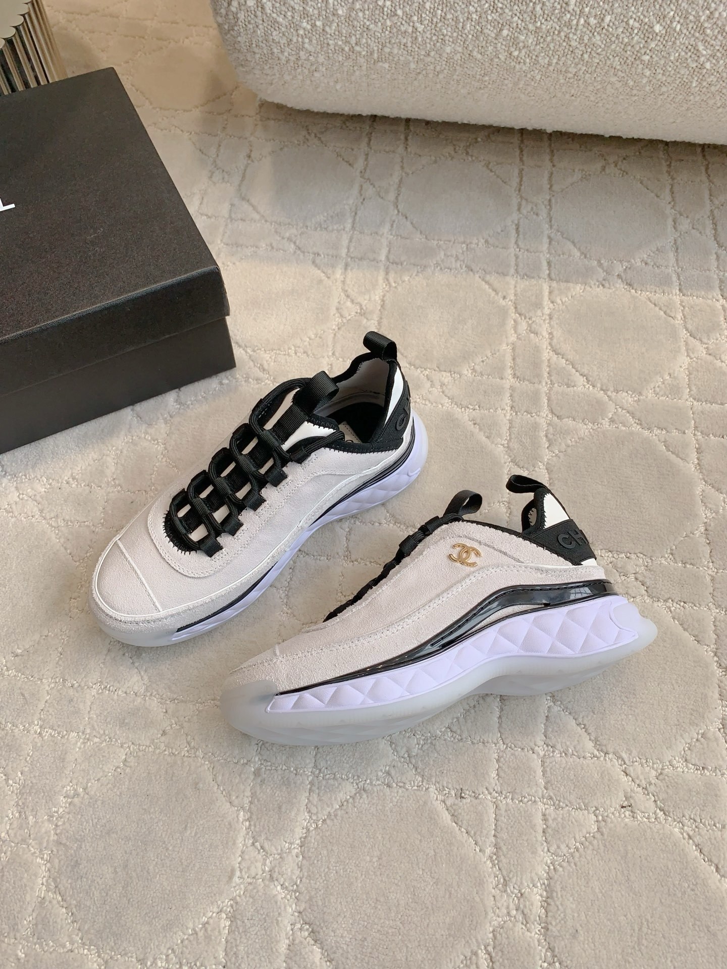 A pair of luxury Chanel white suede sneakers with black accents and gold CC logo placed on a cream quilted carpet. - Hình 4