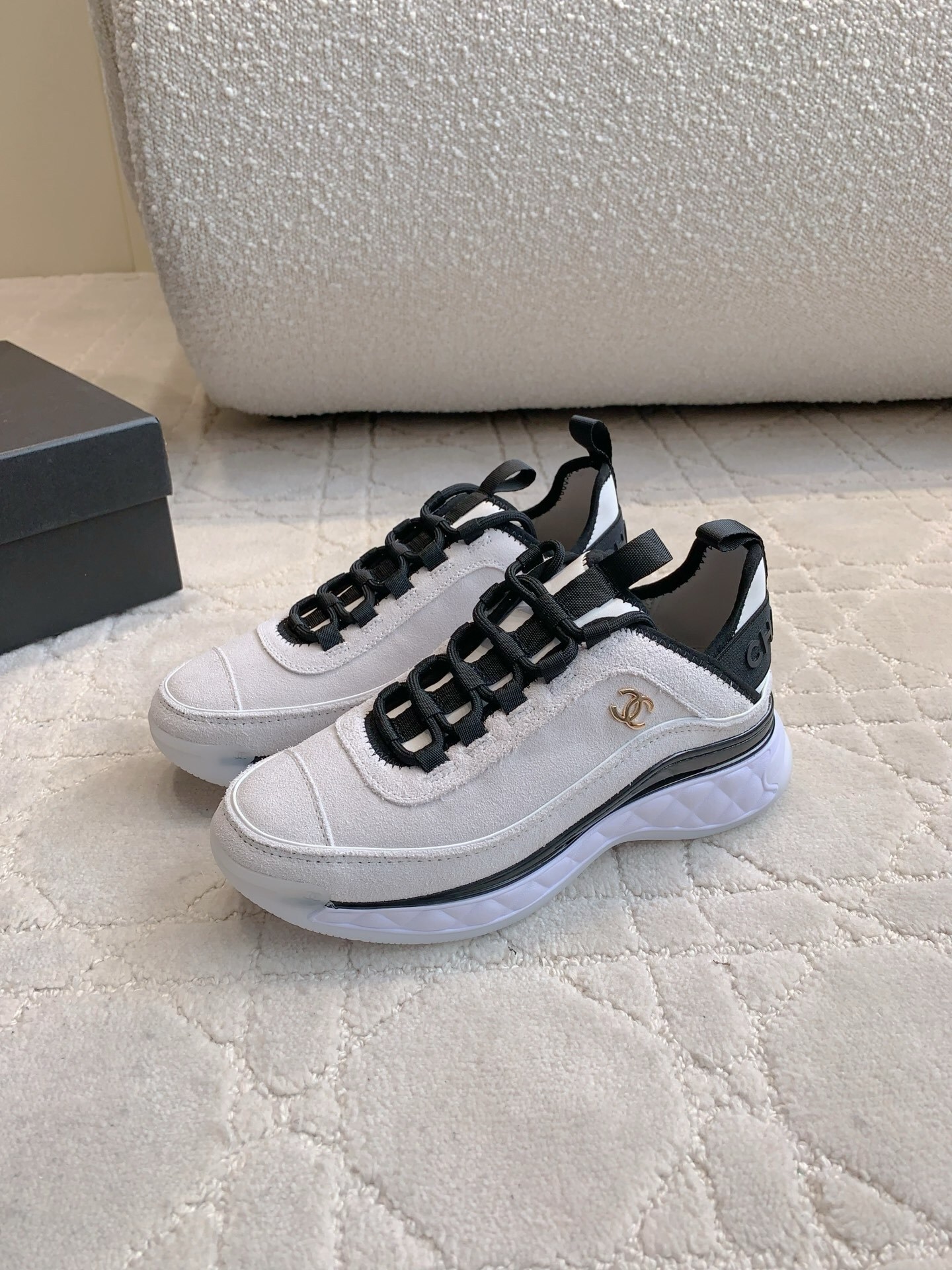 A pair of luxury Chanel white suede sneakers with black accents and gold CC logo placed on a cream quilted carpet. - Hình 2