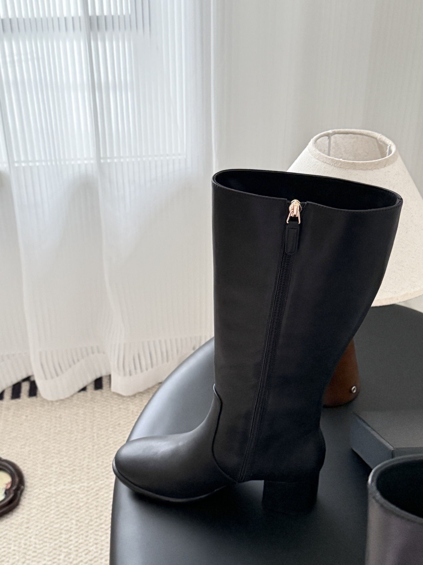 A pair of Chanel black leather knee-high boots with gold CC logo and block heels displayed on a black table next to a Chanel box. - Hình 9