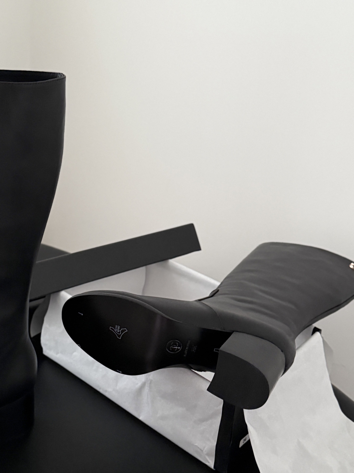 A pair of Chanel black leather knee-high boots with gold CC logo and block heels displayed on a black table next to a Chanel box. - Hình 8