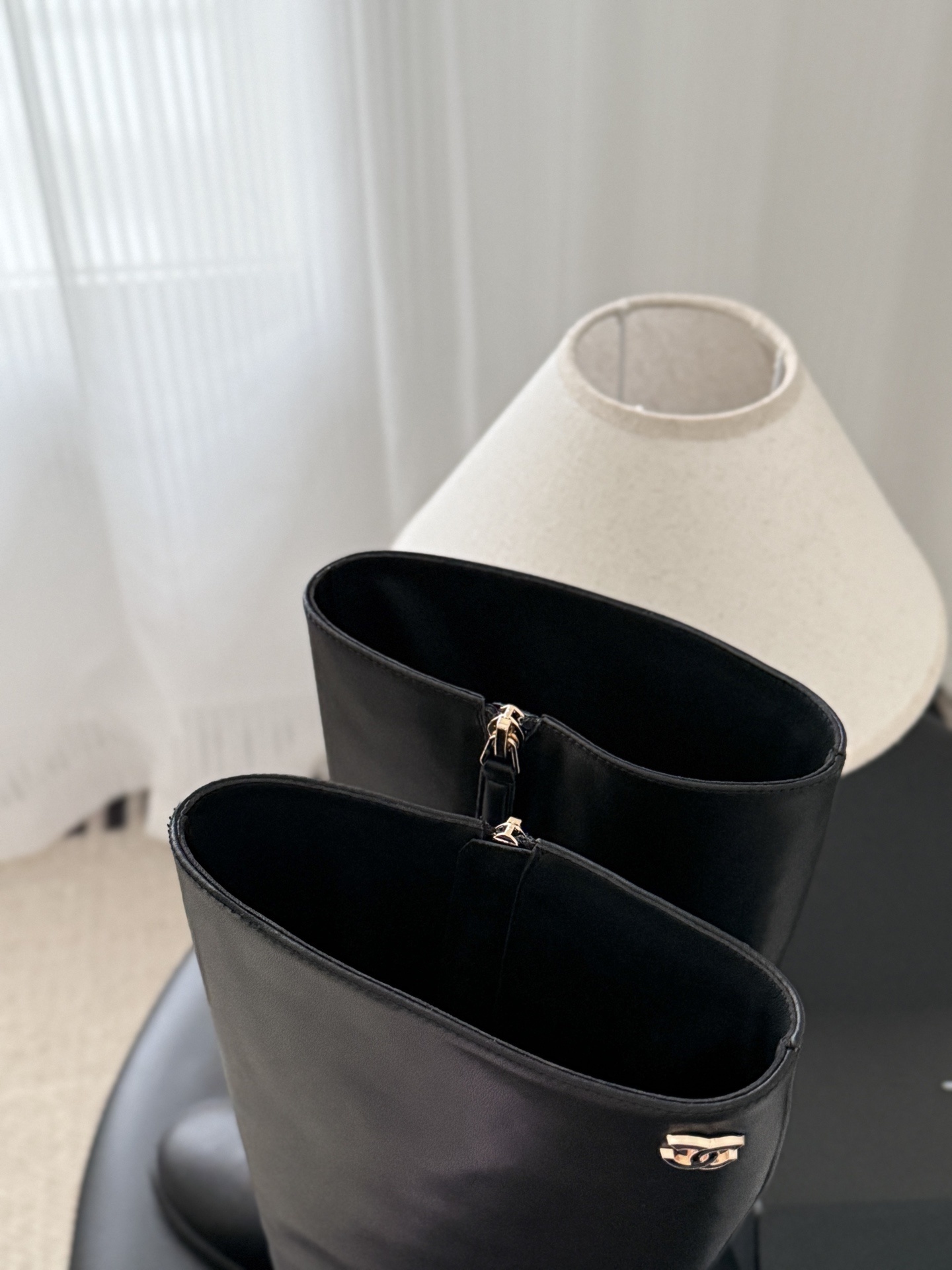 A pair of Chanel black leather knee-high boots with gold CC logo and block heels displayed on a black table next to a Chanel box. - Hình 7