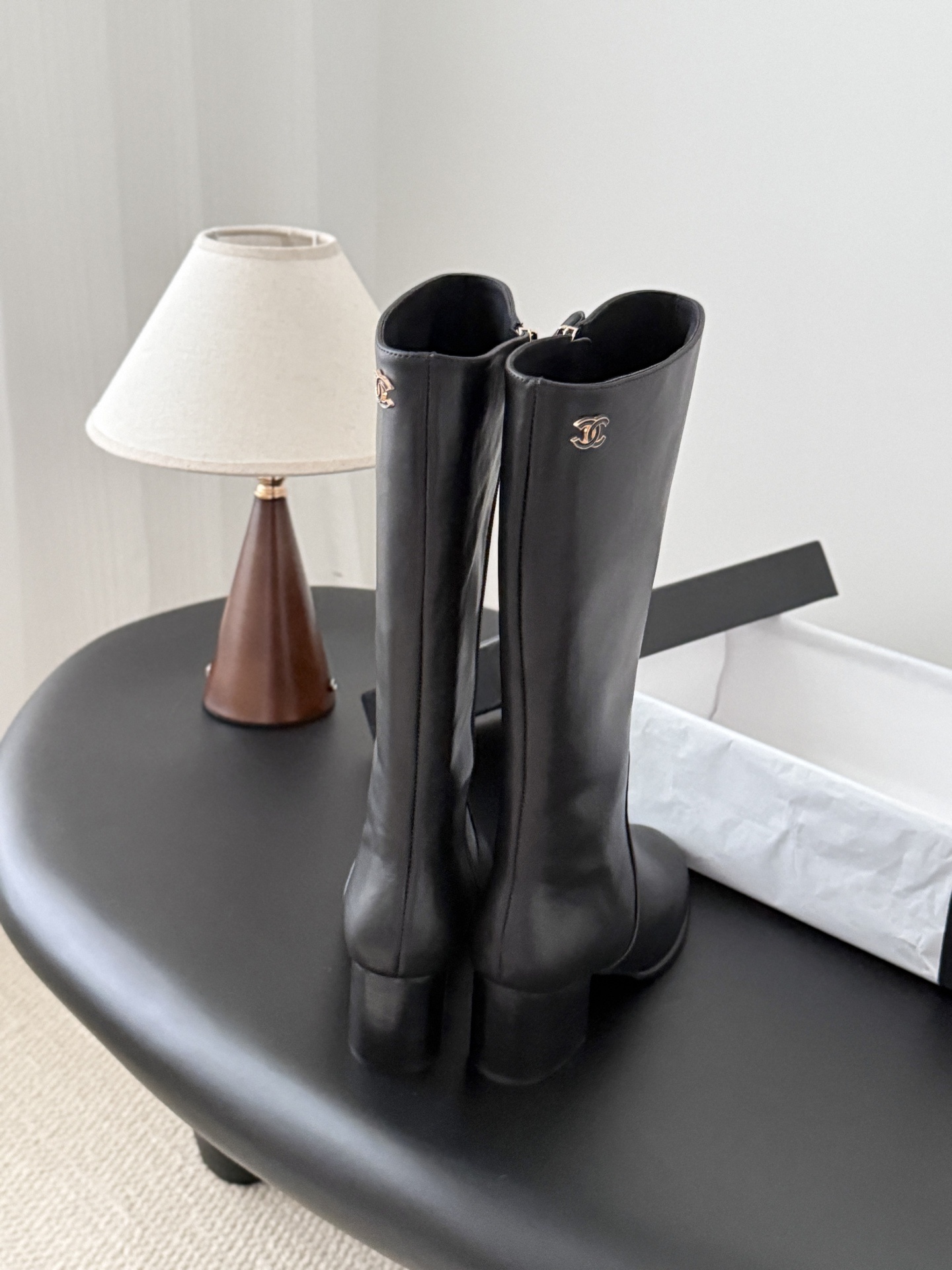 A pair of Chanel black leather knee-high boots with gold CC logo and block heels displayed on a black table next to a Chanel box. - Hình 5