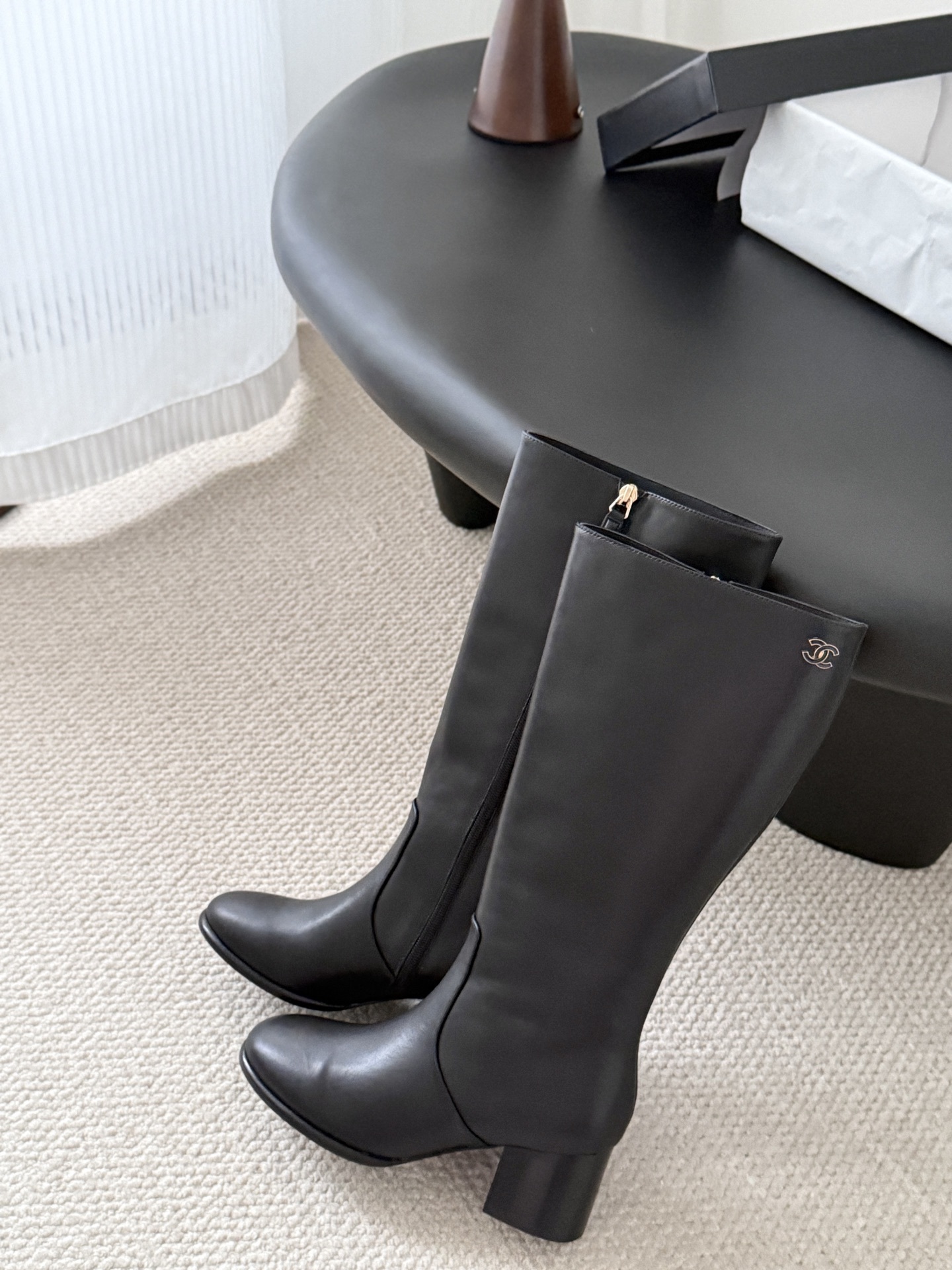 A pair of Chanel black leather knee-high boots with gold CC logo and block heels displayed on a black table next to a Chanel box. - Hình 4