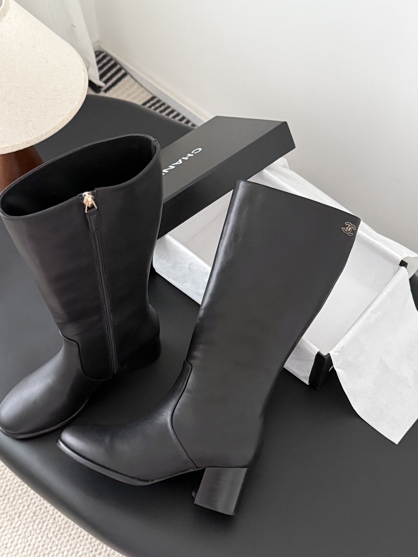 A pair of Chanel black leather knee-high boots with gold CC logo and block heels displayed on a black table next to a Chanel box. - Hình 3