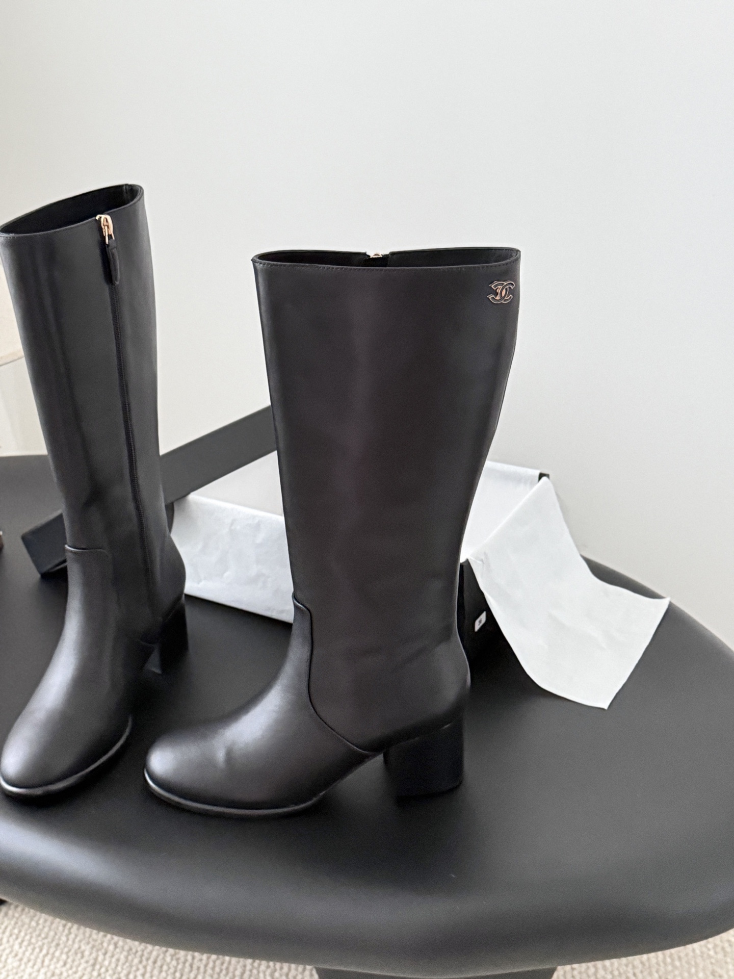 A pair of Chanel black leather knee-high boots with gold CC logo and block heels displayed on a black table next to a Chanel box. - Hình 2