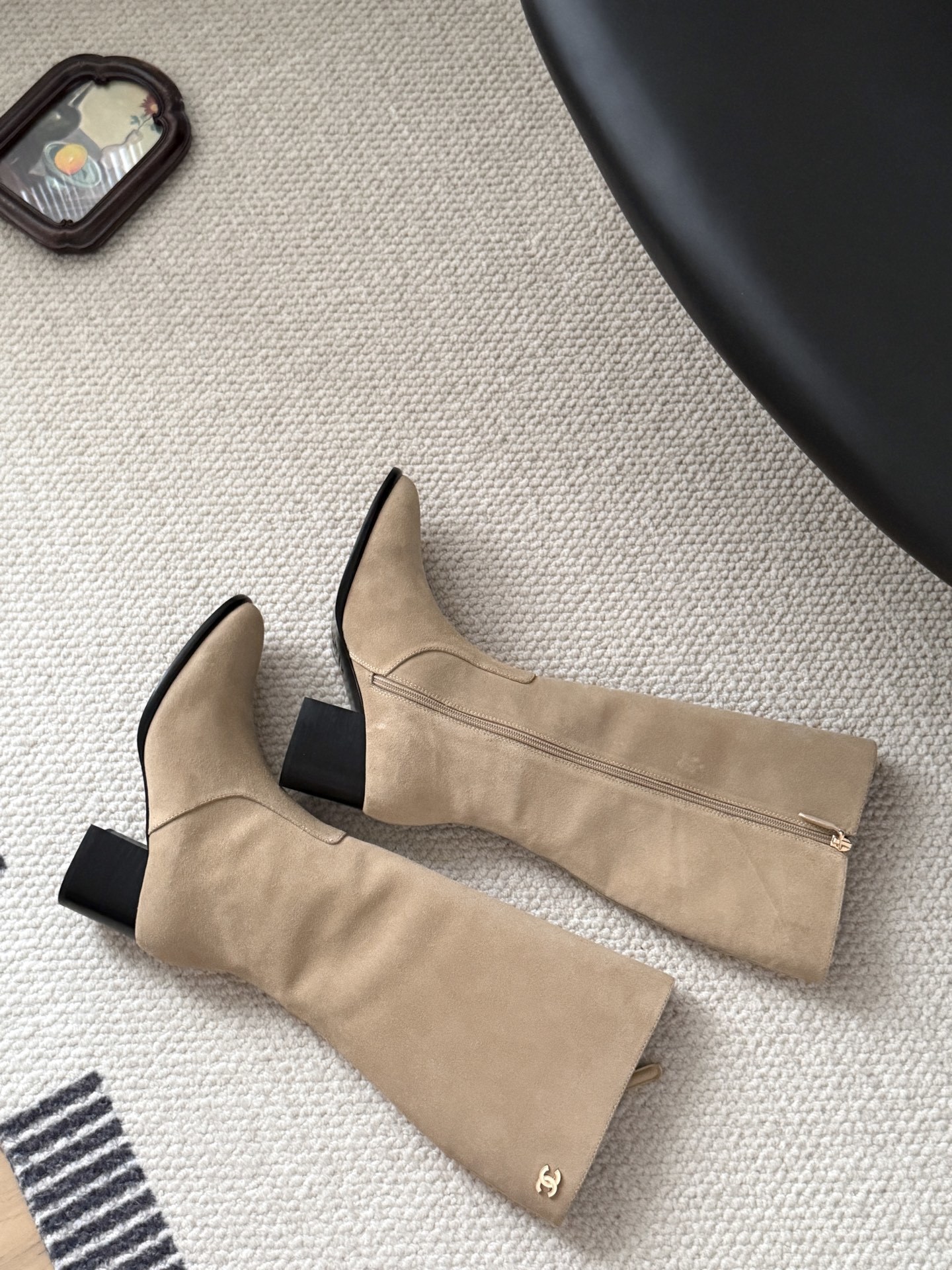 Elegant beige suede knee-high boots with black block heels and gold logo detail on a black table - Hình 9