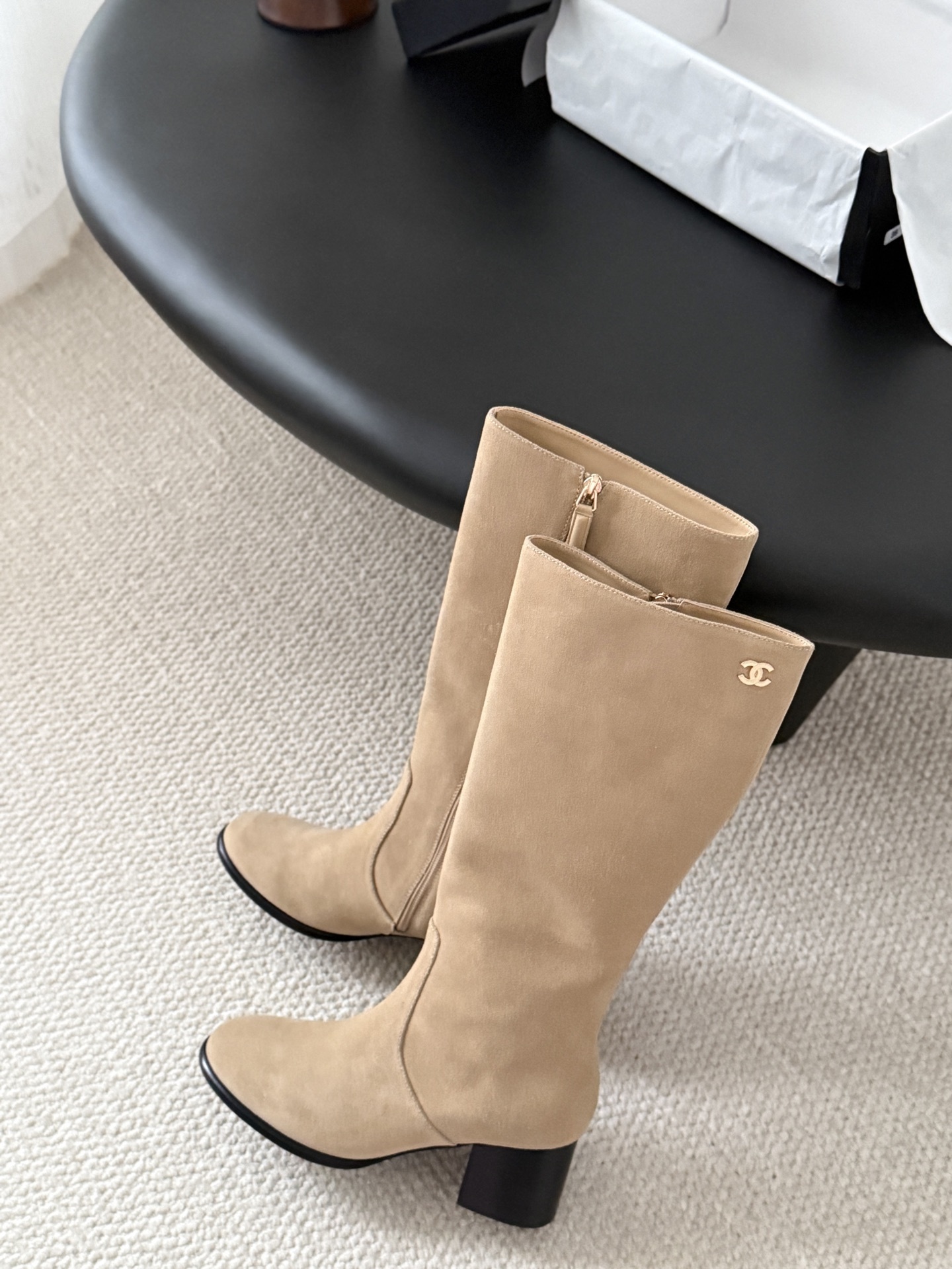 Elegant beige suede knee-high boots with black block heels and gold logo detail on a black table - Hình 8
