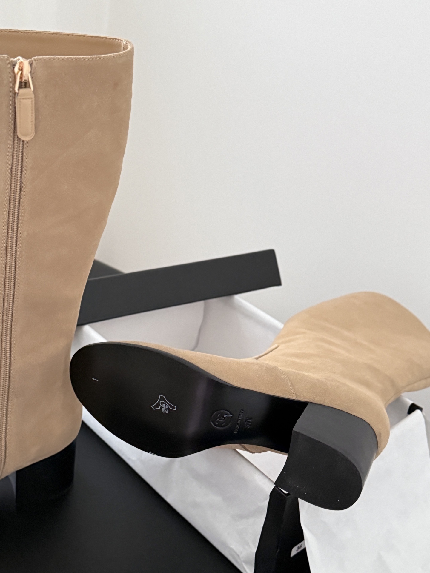 Elegant beige suede knee-high boots with black block heels and gold logo detail on a black table - Hình 6