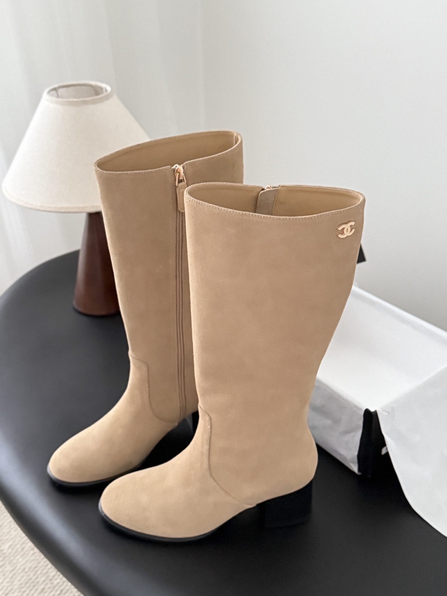 Elegant beige suede knee-high boots with black block heels and gold logo detail on a black table - Hình 5