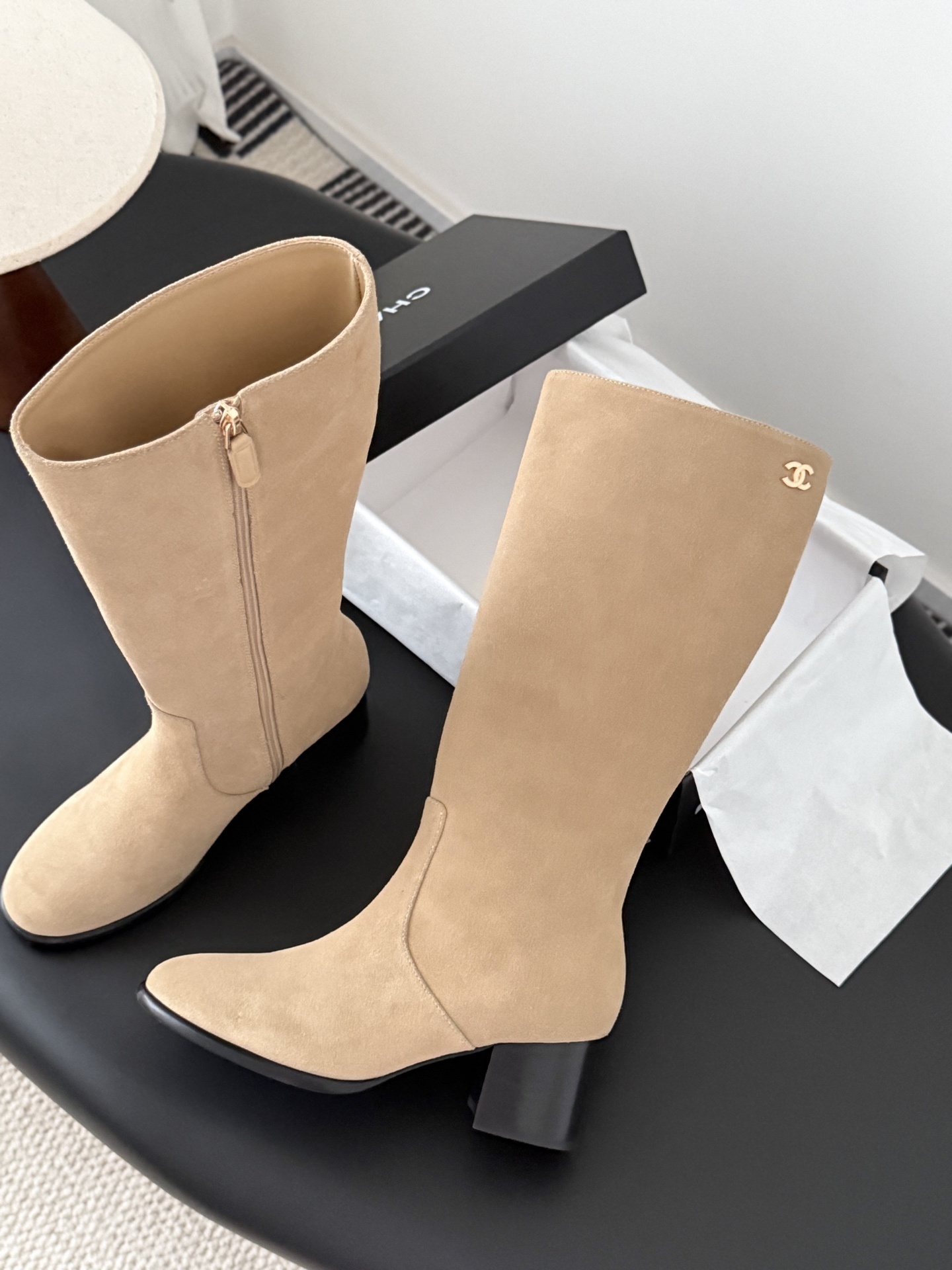 Elegant beige suede knee-high boots with black block heels and gold logo detail on a black table - Hình 3