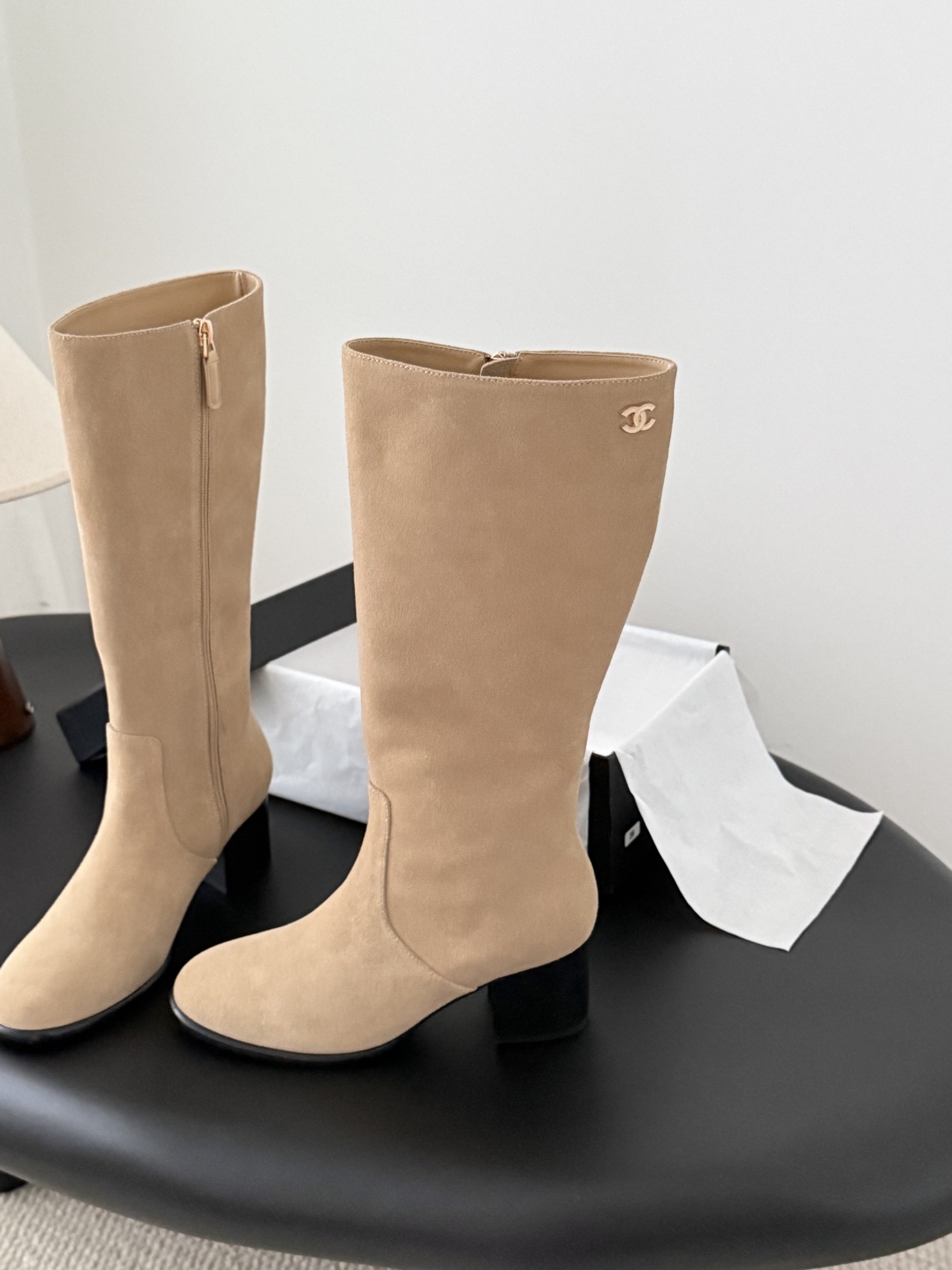 Elegant beige suede knee-high boots with black block heels and gold logo detail on a black table - Hình 2