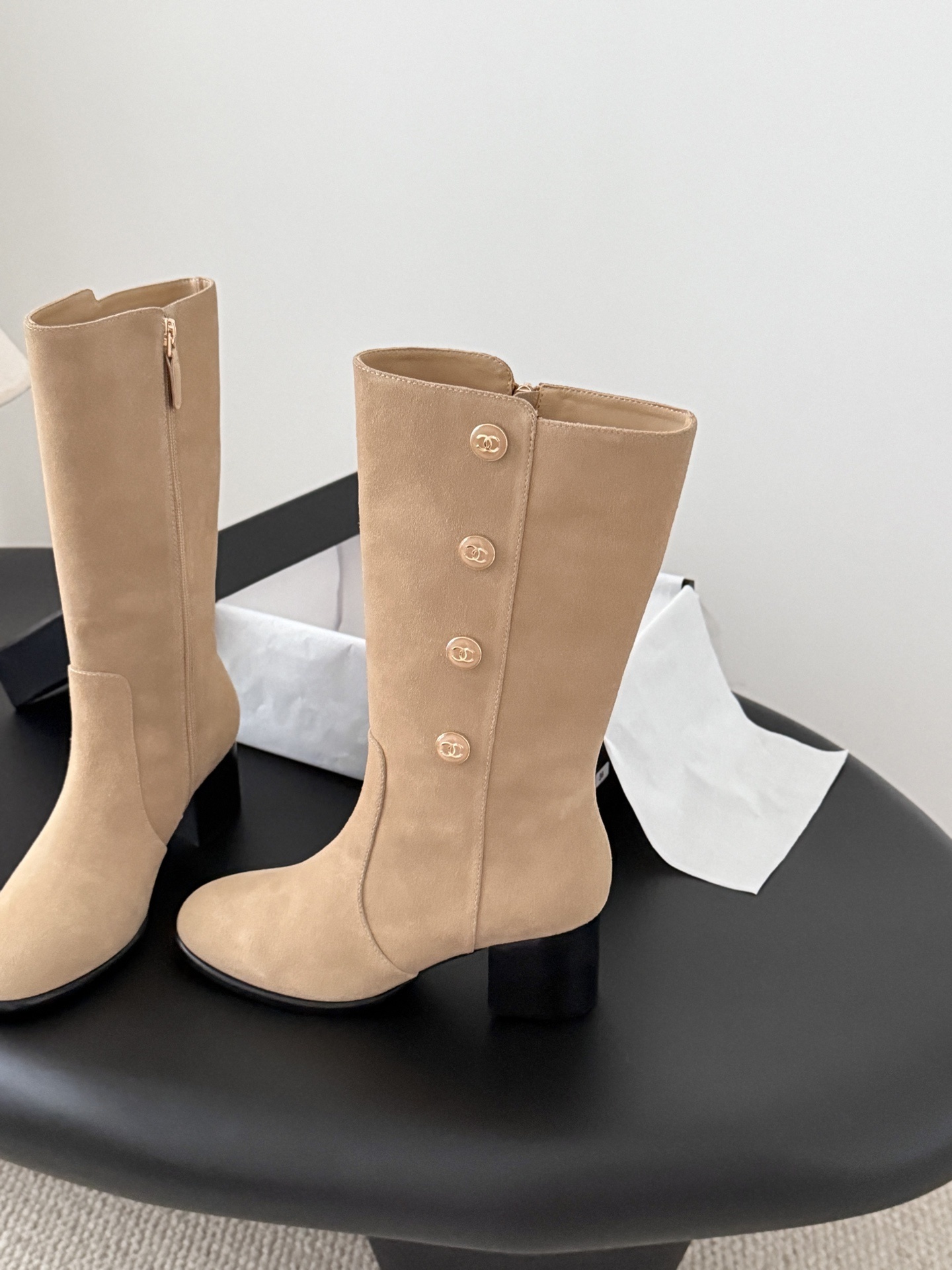 Pair of Chanel beige suede mid-calf boots with gold CC logo buttons and black block heels on a black table. - Hình 3