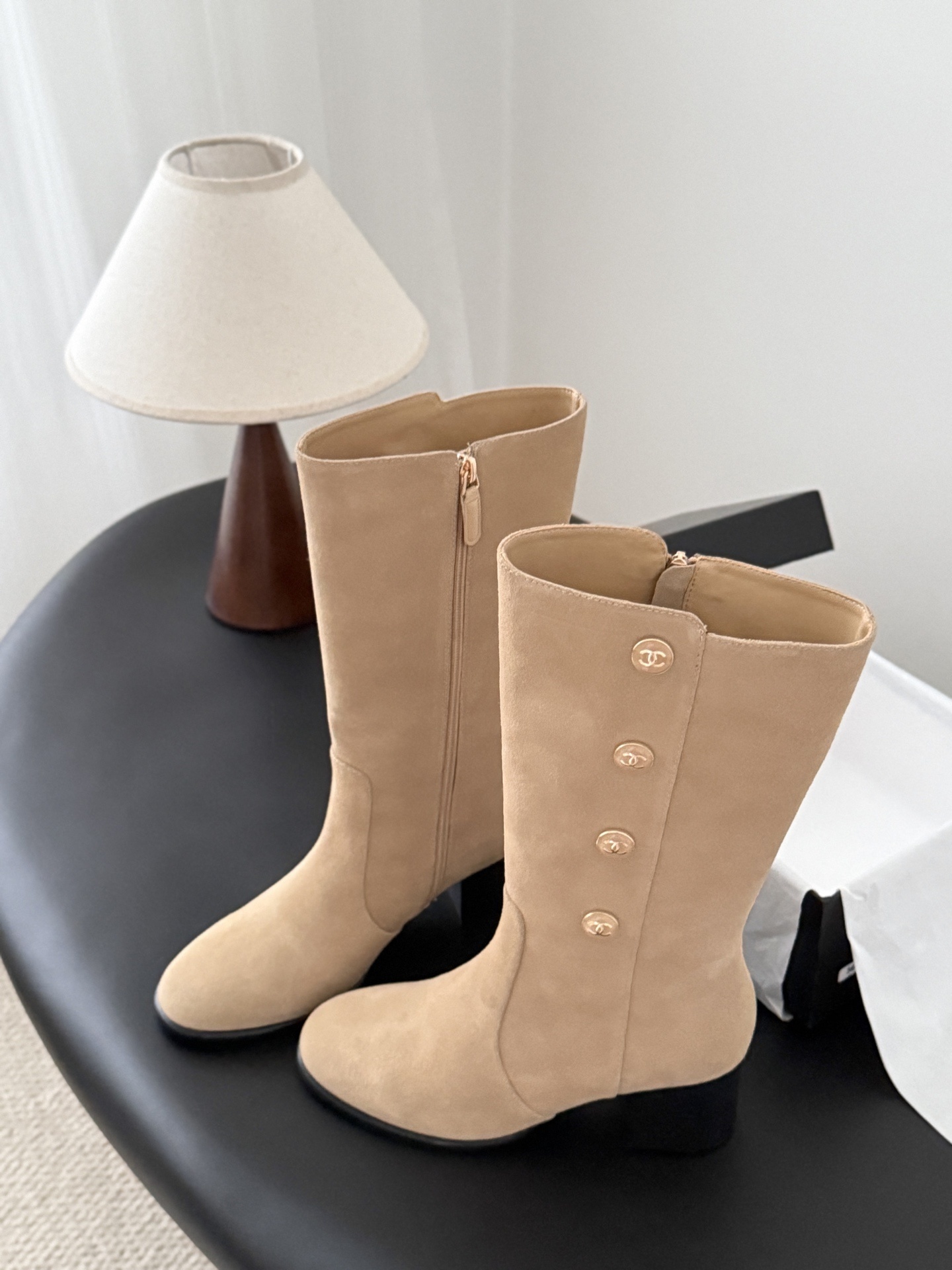 Pair of Chanel beige suede mid-calf boots with gold CC logo buttons and black block heels on a black table. - Hình 2