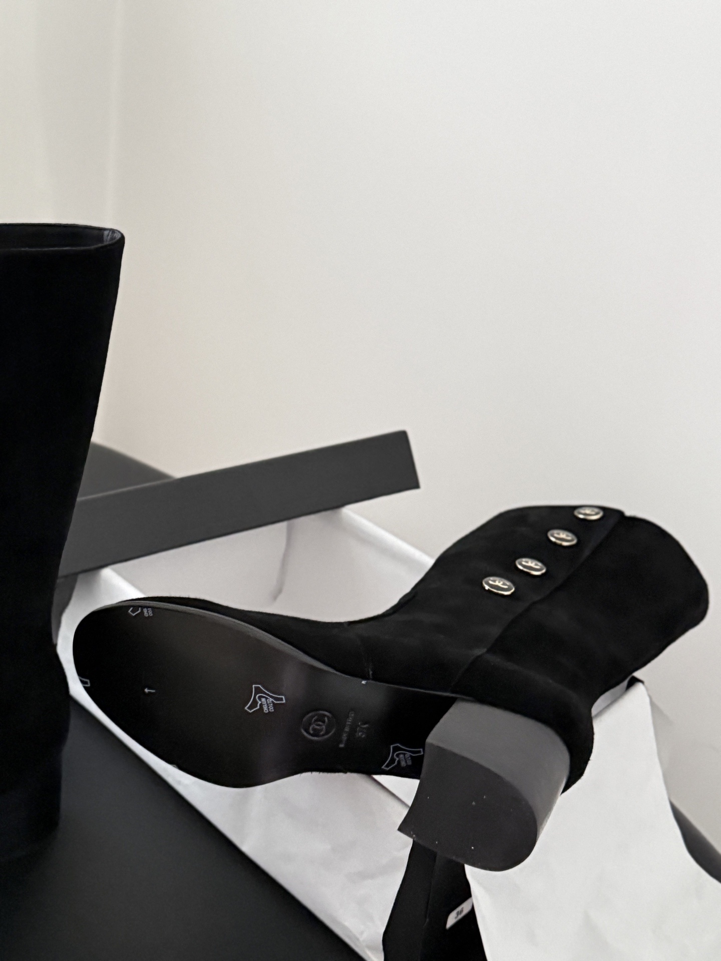 A pair of elegant black suede mid-calf boots with decorative gold logo buttons and a block heel, displayed next to a designer shoe box. - Hình 4