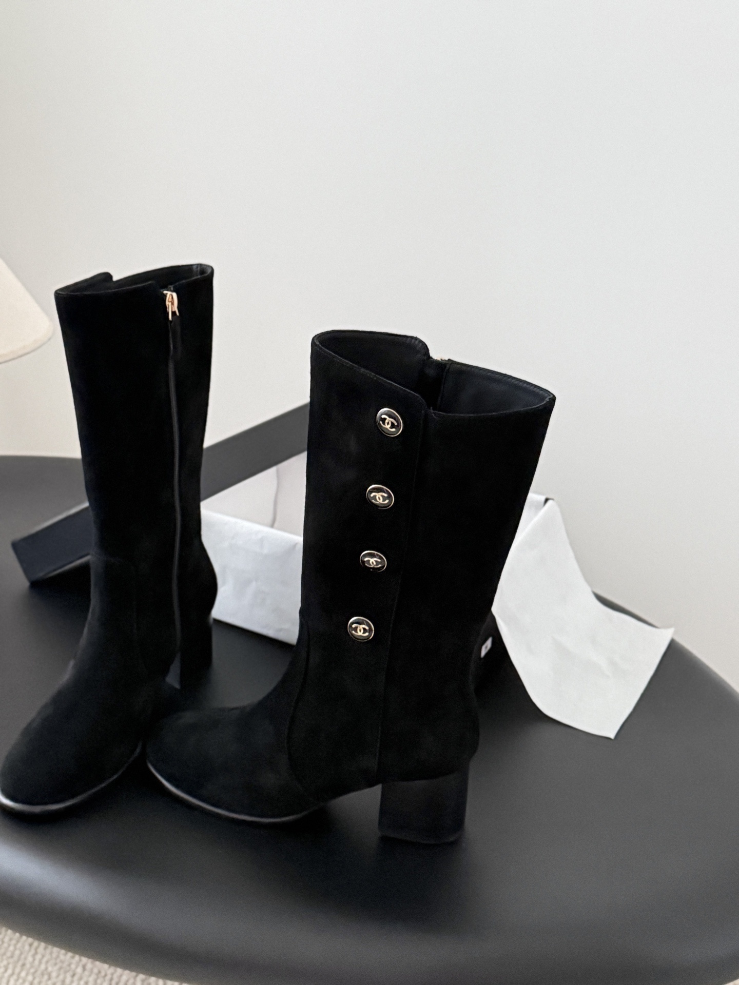 A pair of elegant black suede mid-calf boots with decorative gold logo buttons and a block heel, displayed next to a designer shoe box. - Hình 2