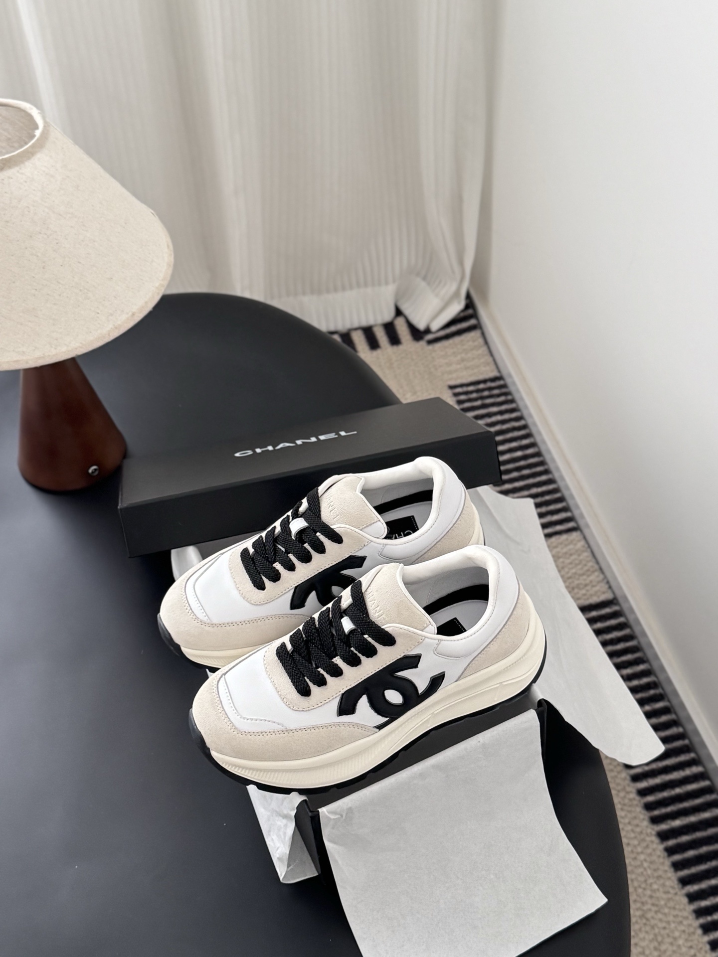A pair of Chanel white and beige suede sneakers with black CC logo and laces displayed next to a black Chanel shoe box. - Hình 8