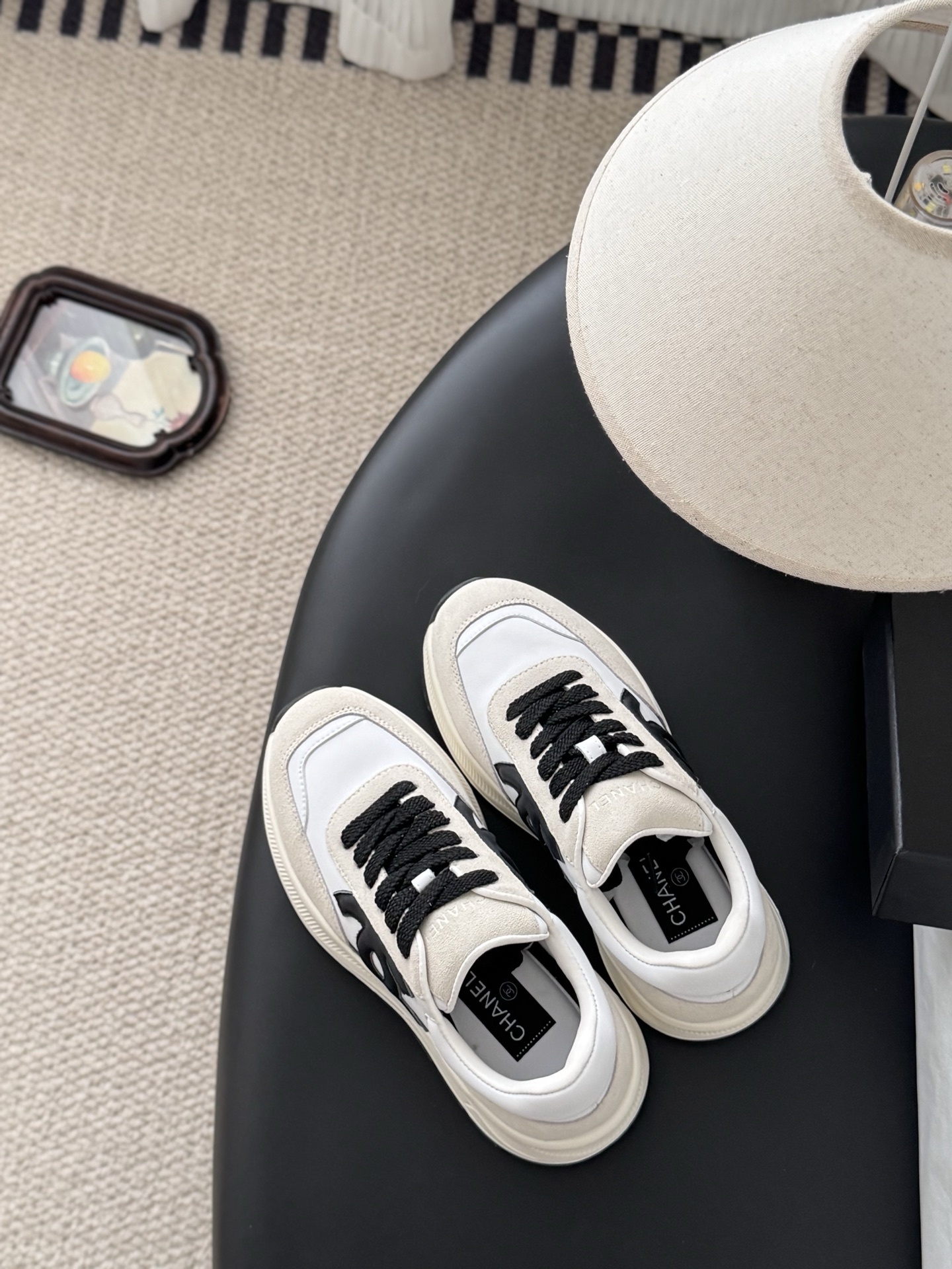 A pair of Chanel white and beige suede sneakers with black CC logo and laces displayed next to a black Chanel shoe box. - Hình 7